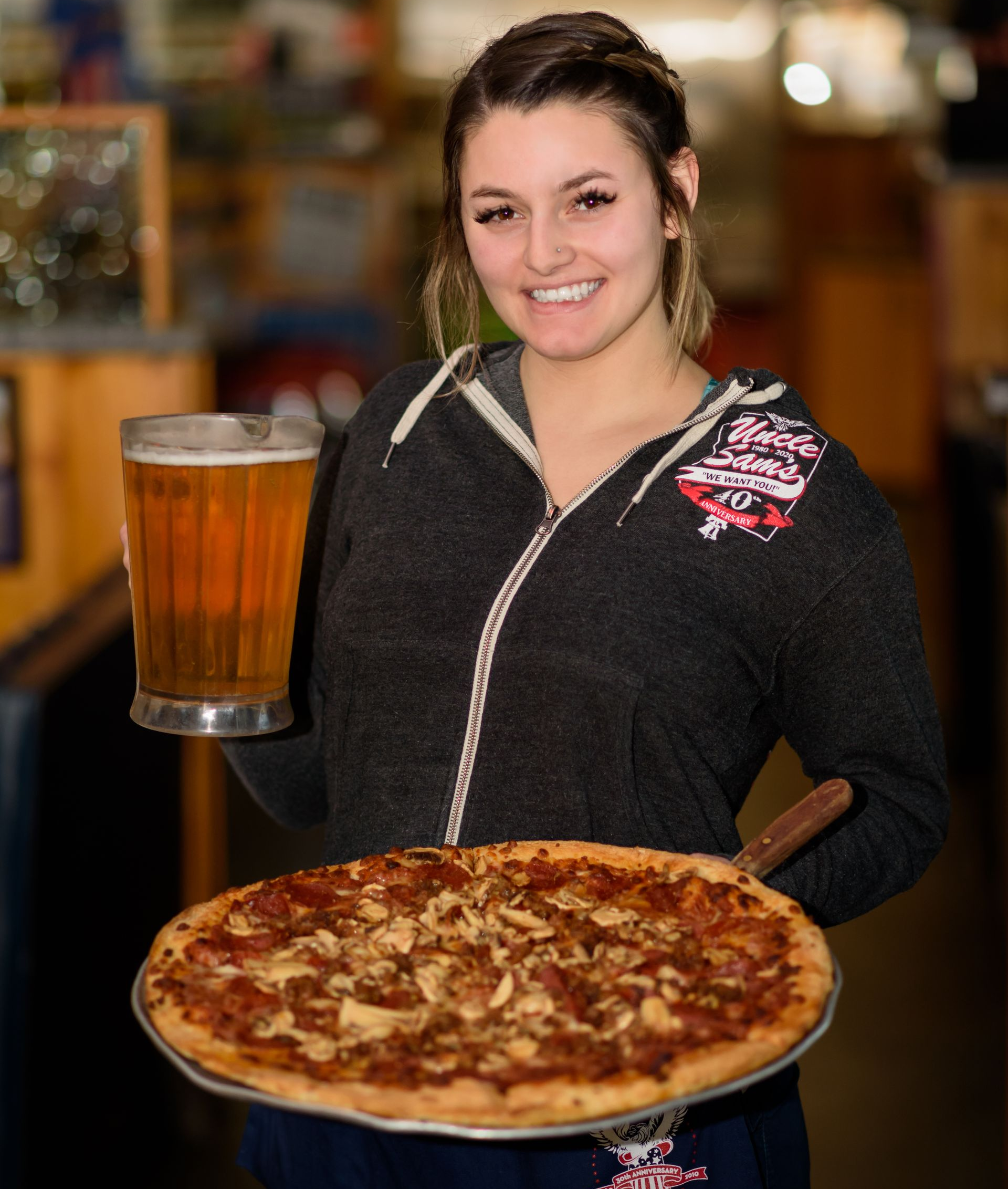 Smiling server holding a beer and large pizza in a restaurant