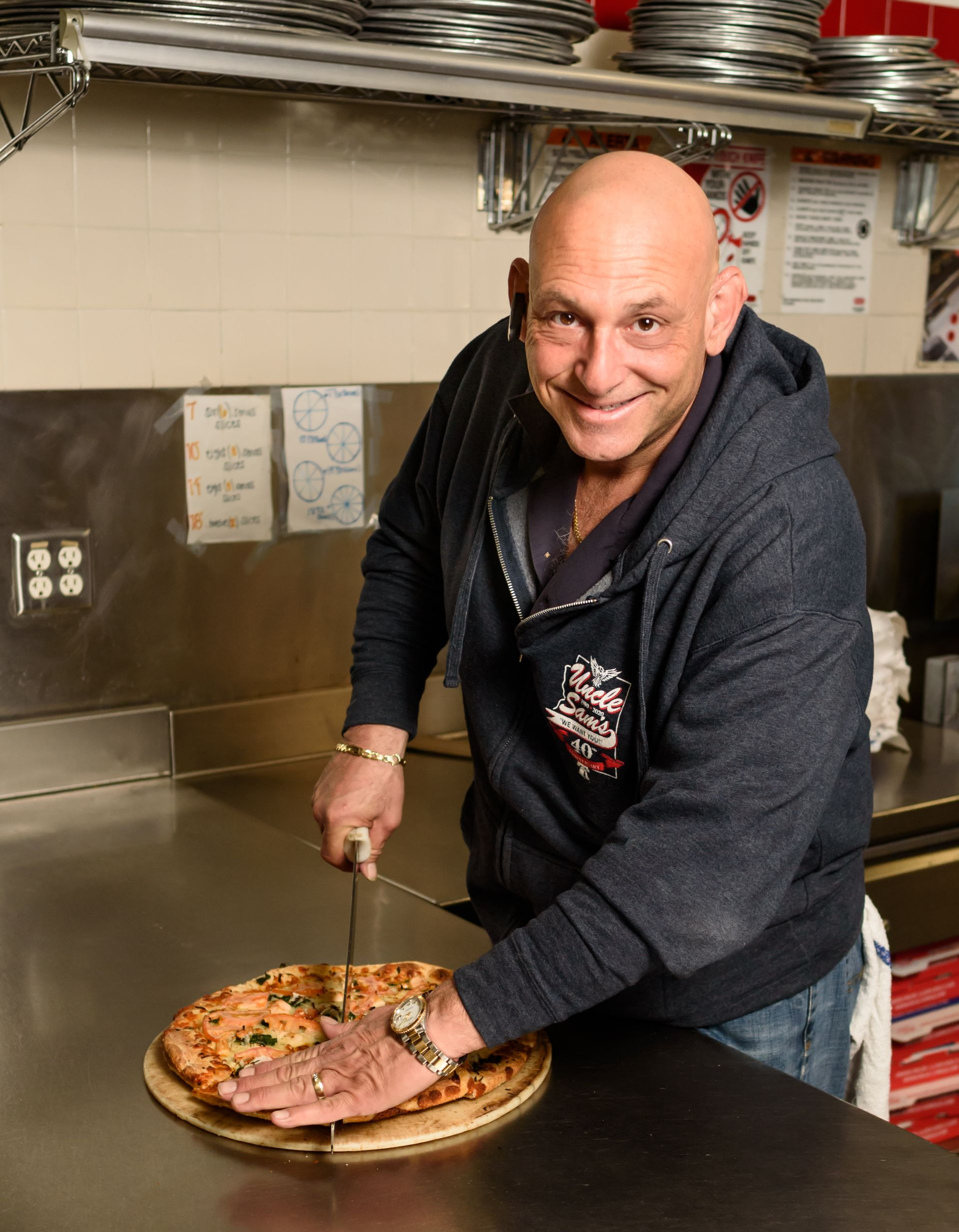 Man smiling in a kitchen, cutting a pizza on a counter with a large knife.