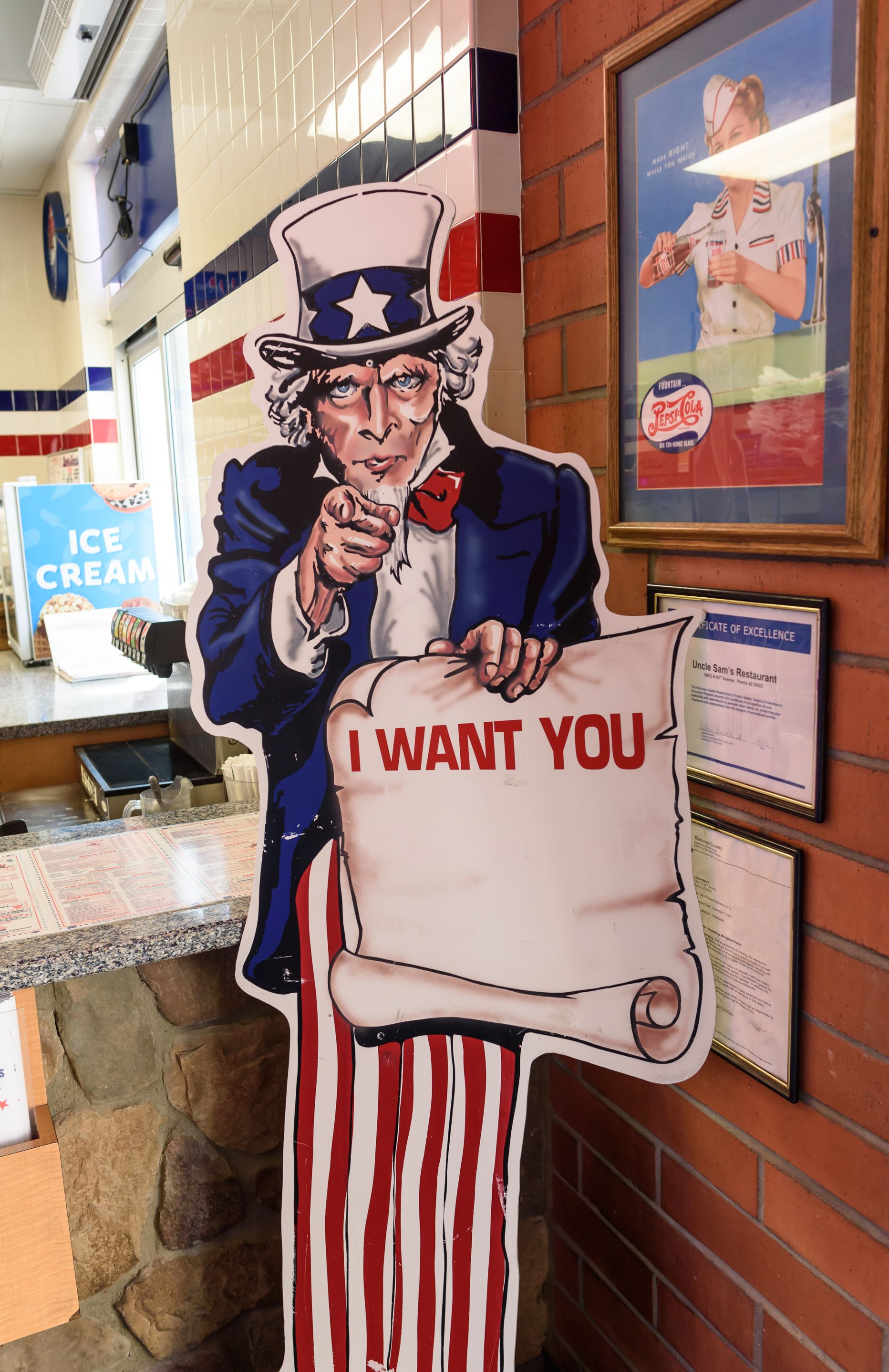 Uncle Sam cutout holding “I WANT YOU” sign beside brick wall and ice cream shop window