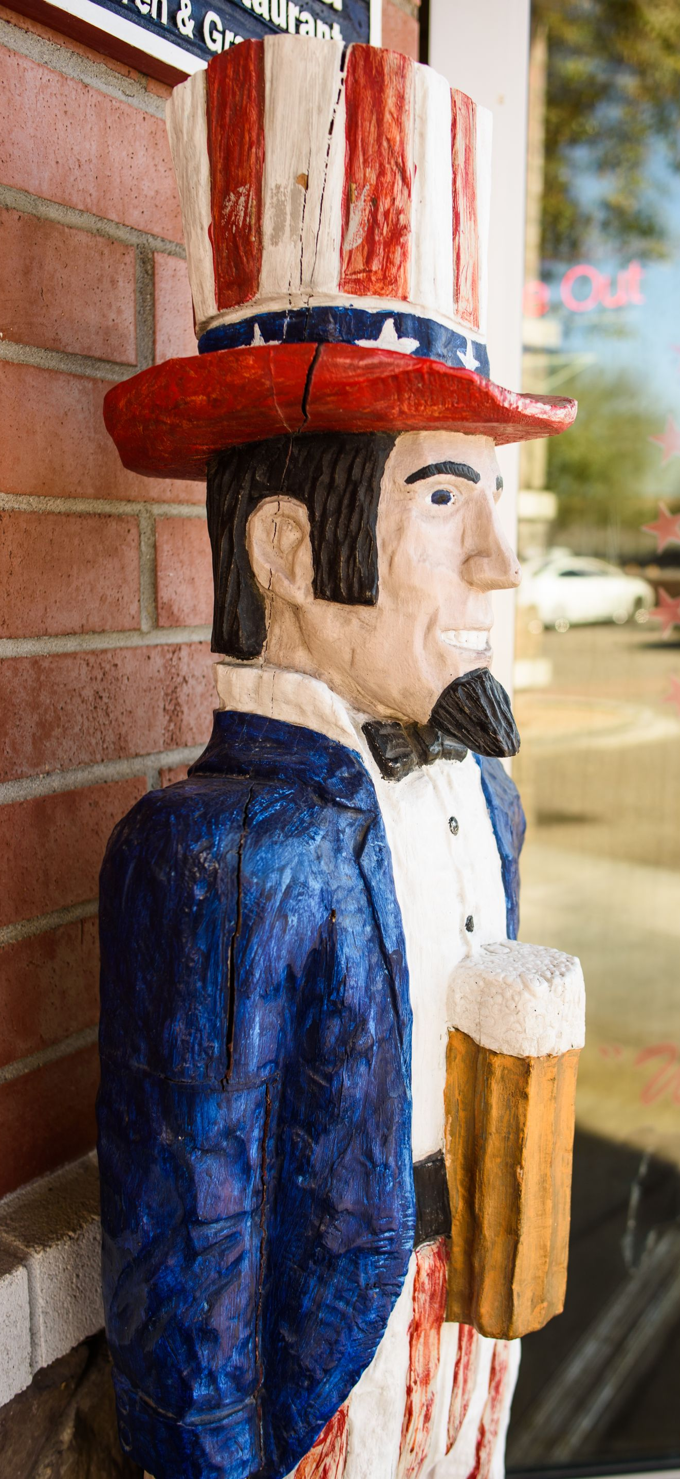 Wooden Uncle Sam statue in red, white, and blue leaning by a brick wall and storefront window