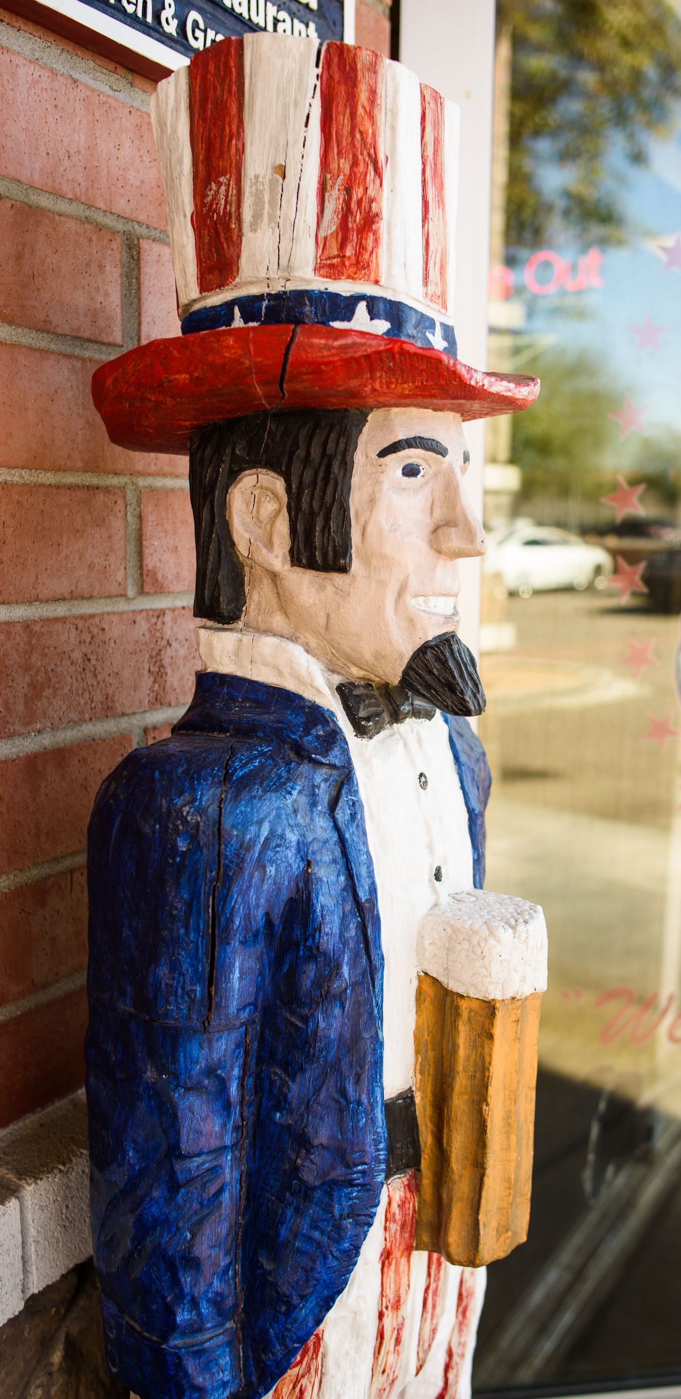 Painted wooden statue of a man in a red-white-blue hat and blue coat by a brick wall.
