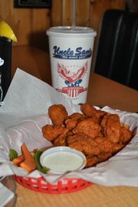 A basket of breaded fried chicken pieces and dipping sauce next to a paper cup from Uncle Sam's on a wooden table.