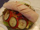A submarine sandwich with sliced deli meat, cucumbers, tomatoes, and greens on a bed of shredded lettuce in a container.