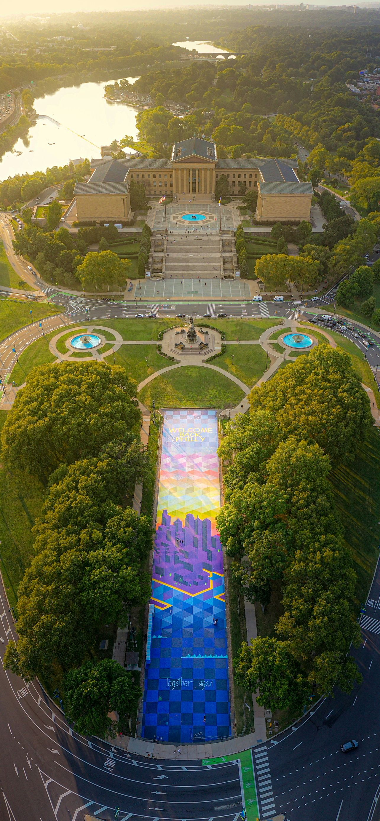 Aerial view of a city park with a colorful rainbow walkway, fountains, and a large building at sunset.