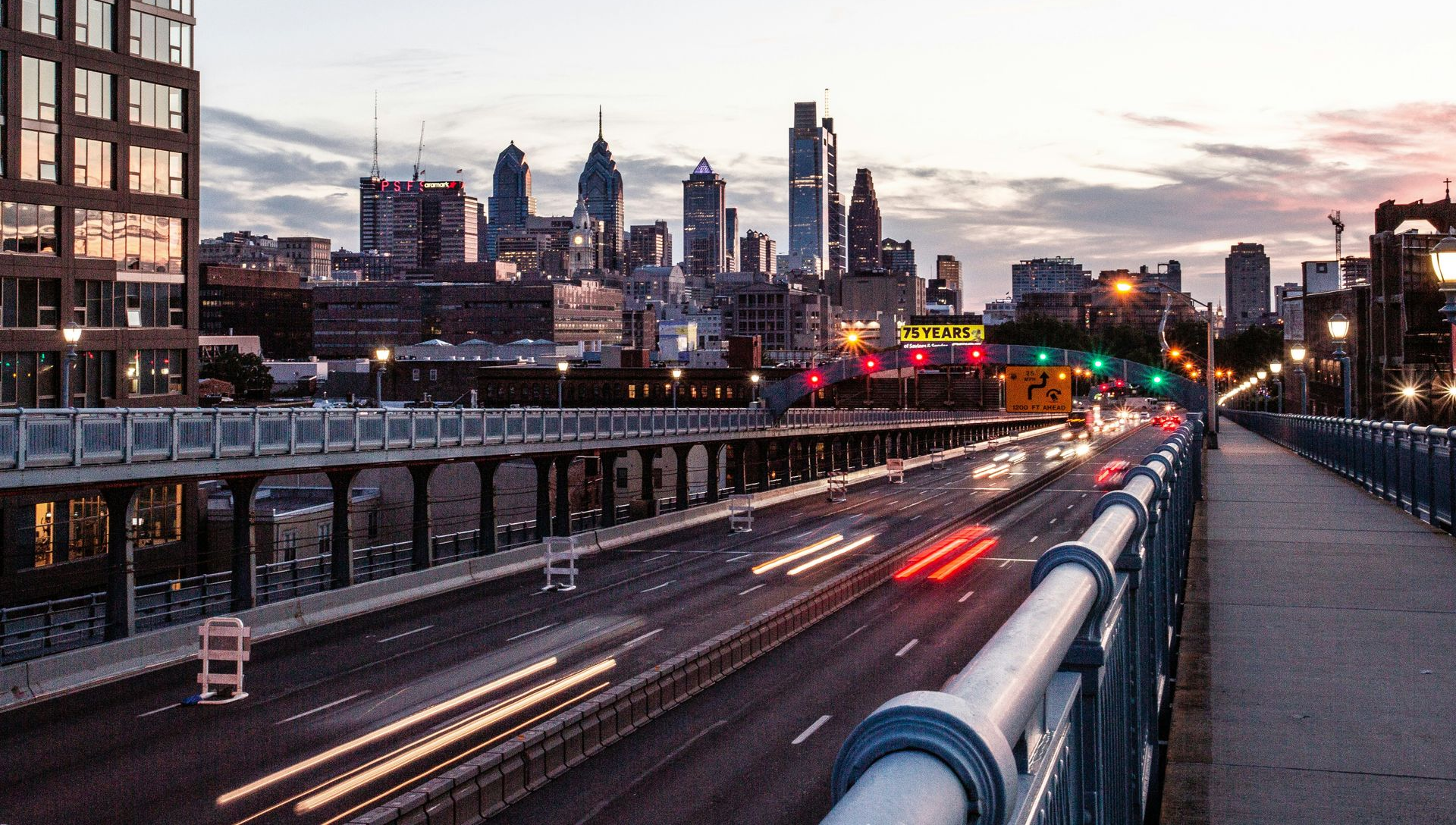 City skyline at dusk above a busy highway, with light trails and a pedestrian bridge railing in the foreground