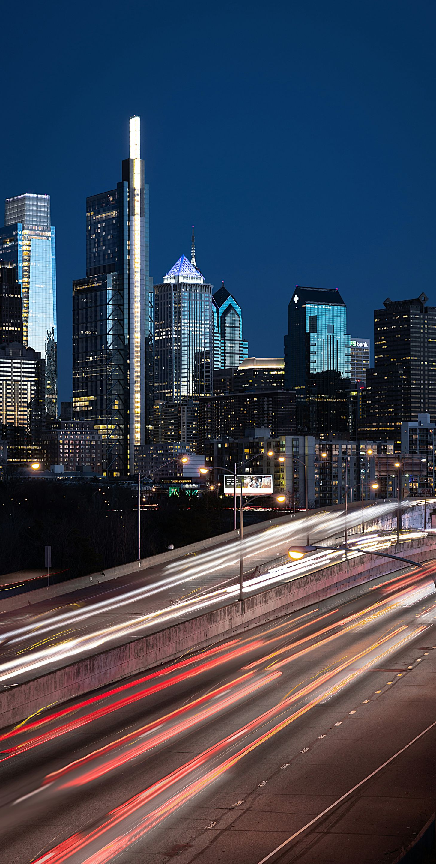 A long-exposure shot of Philadelphia's skyline at night, with light trails from highway traffic in the foreground.