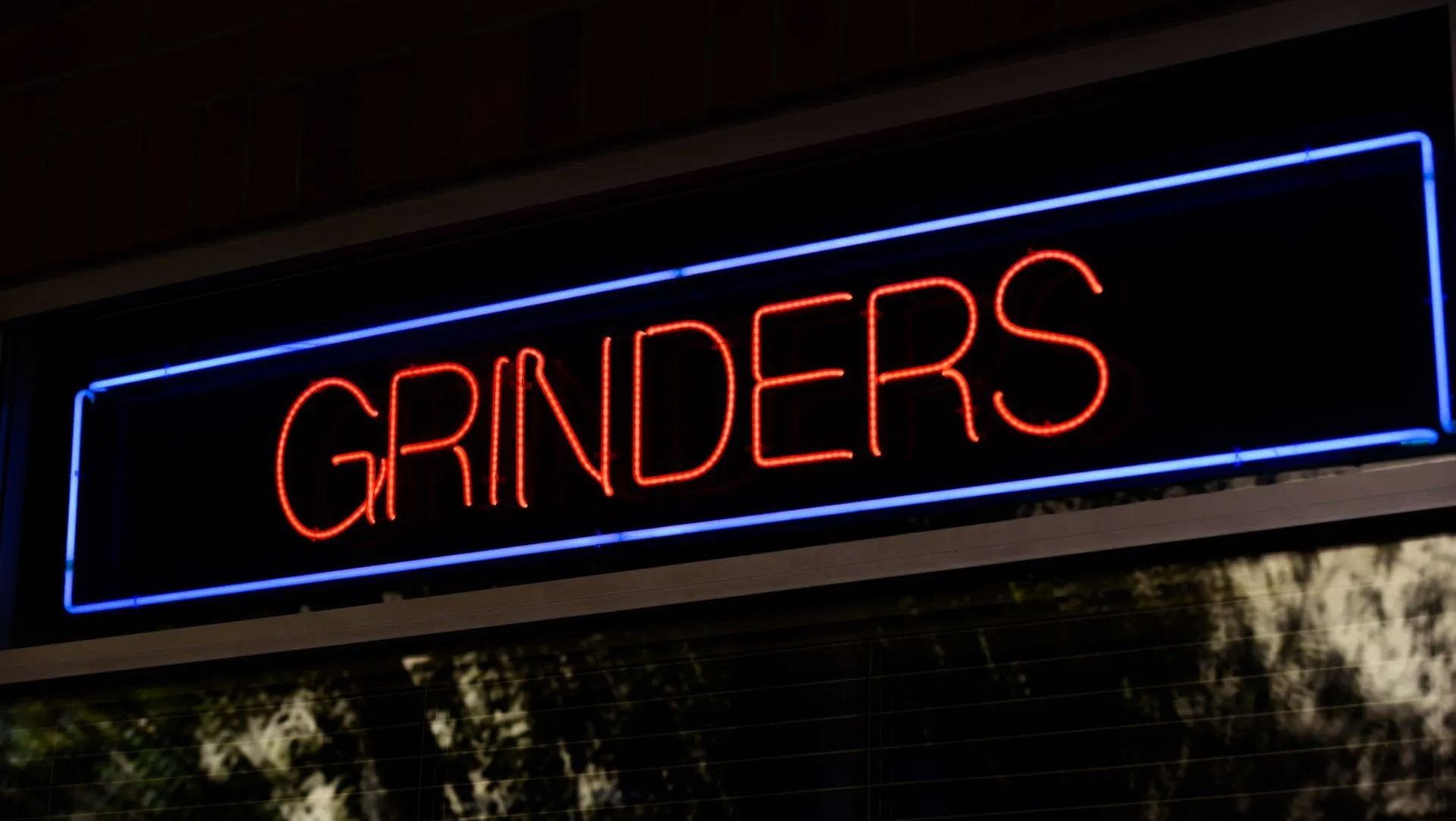 Neon “GRINDERS” sign in red inside a blue-lit rectangular frame at night