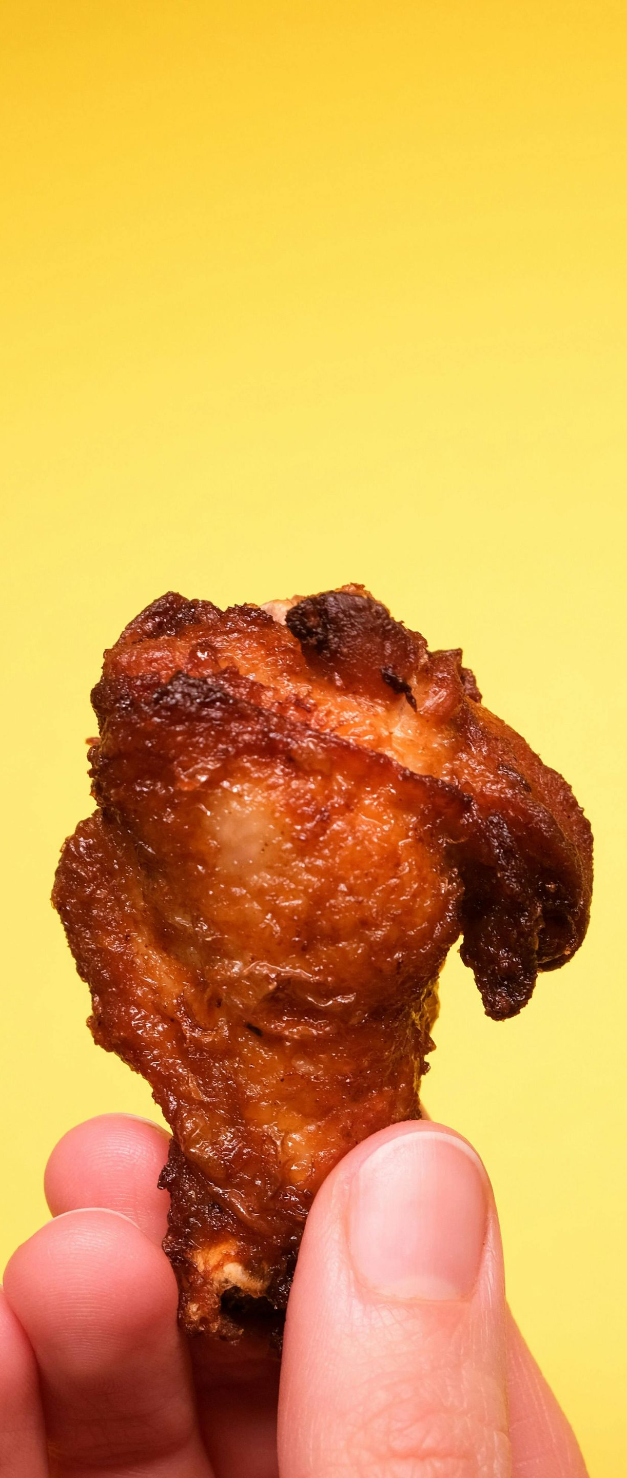 A hand holding a fried chicken drumstick against a bright yellow background