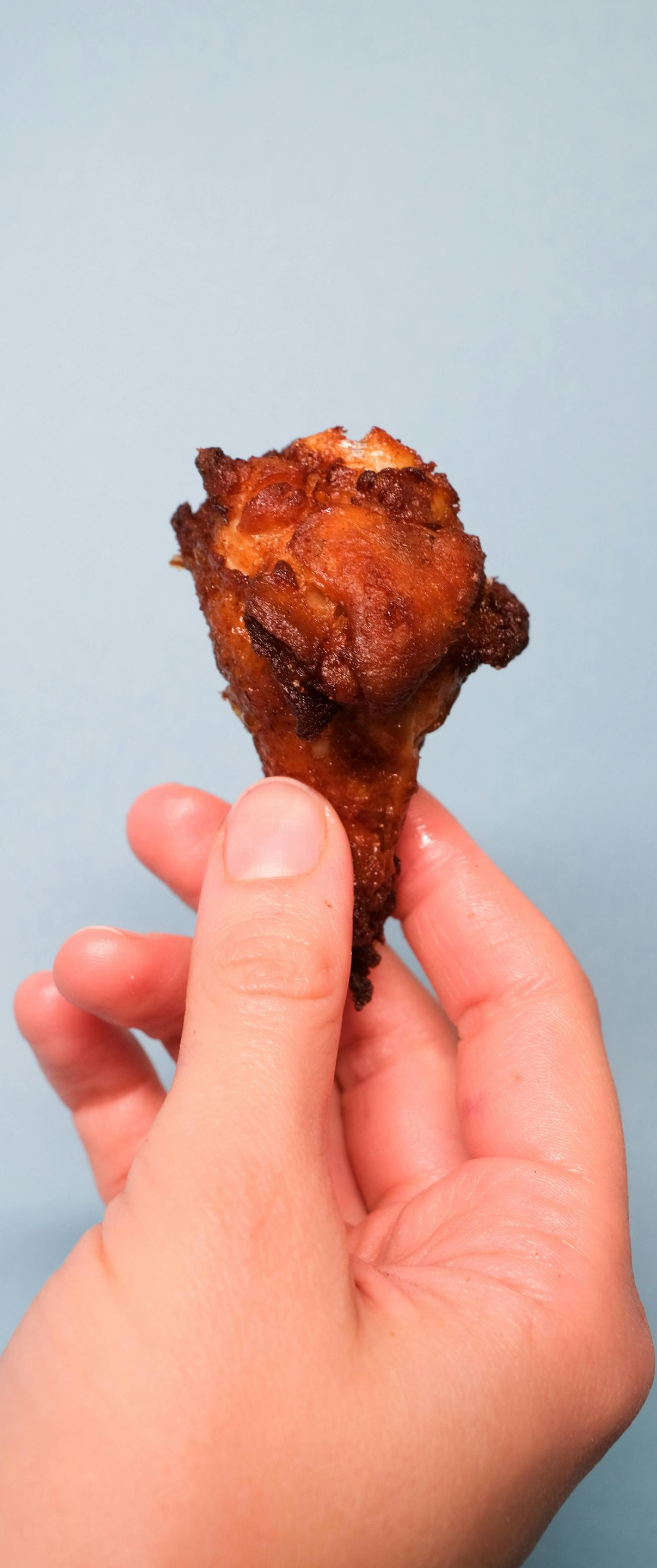 Hand holding a crispy, browned chicken drumstick against a light blue background