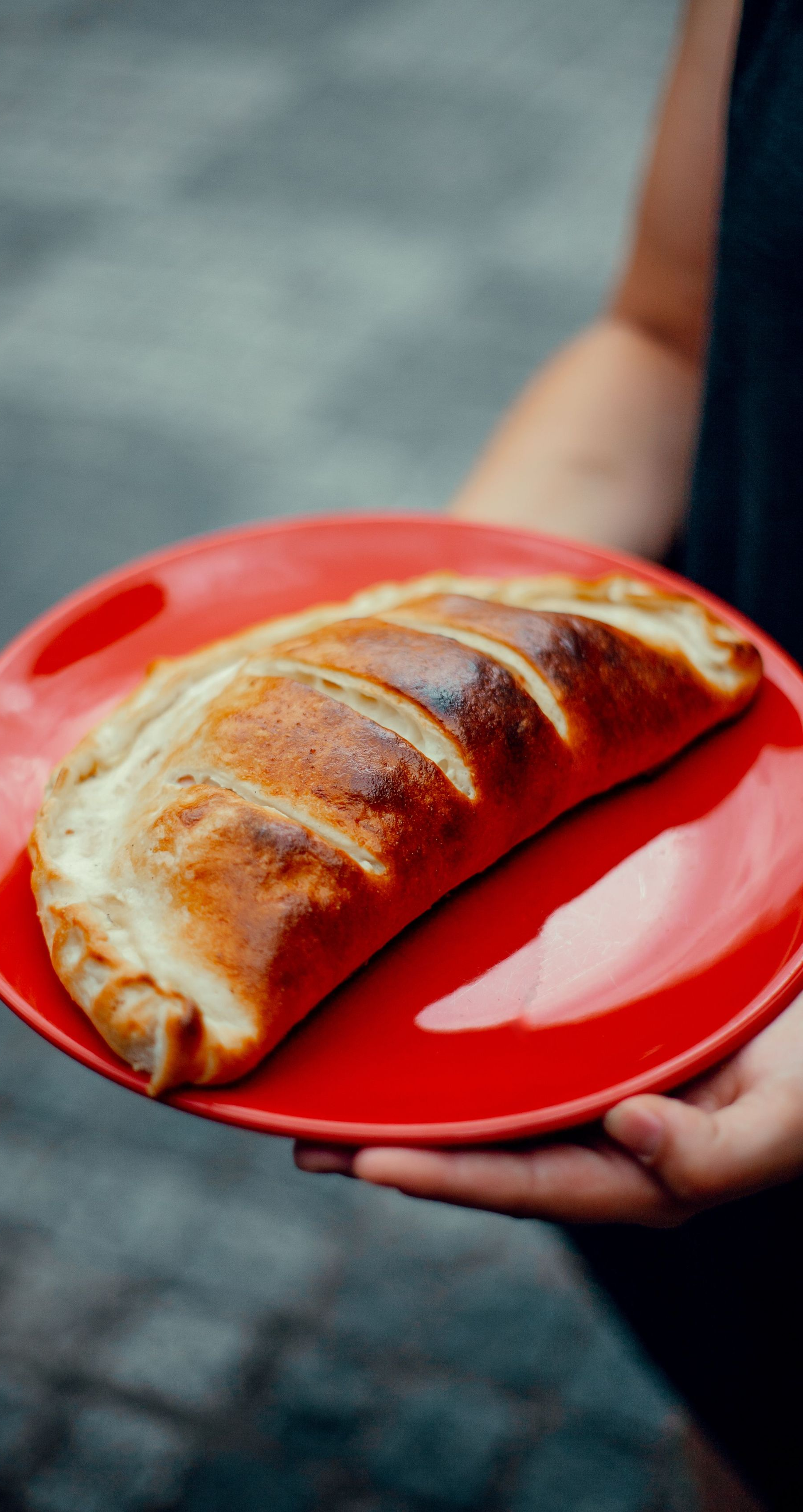 A red plate holding a baked pastry, being carried in someone’s hands outdoors.