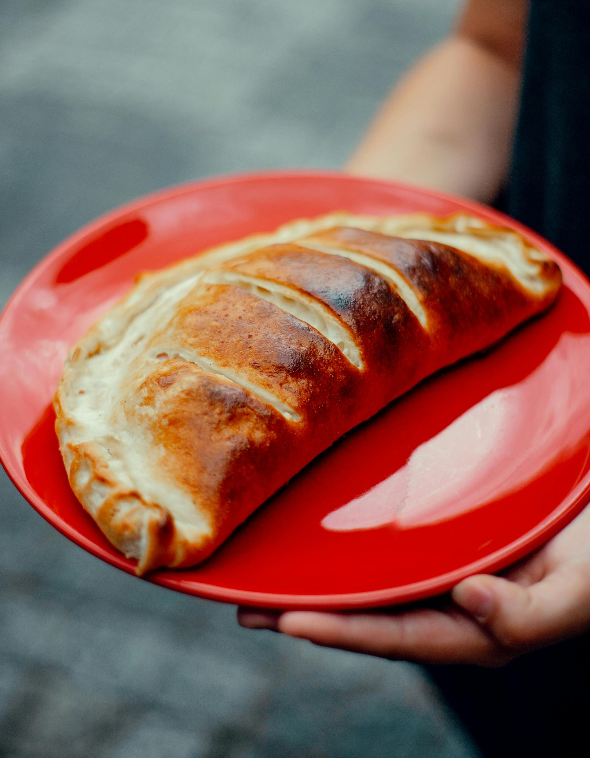 A baked pastry on a red plate being held outdoors by a person’s hands.