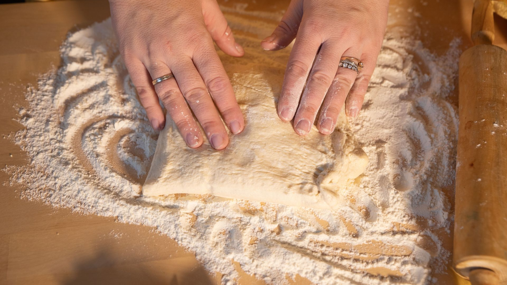Hands kneading flour dough on a wooden surface, with flour scattered around the workspace.