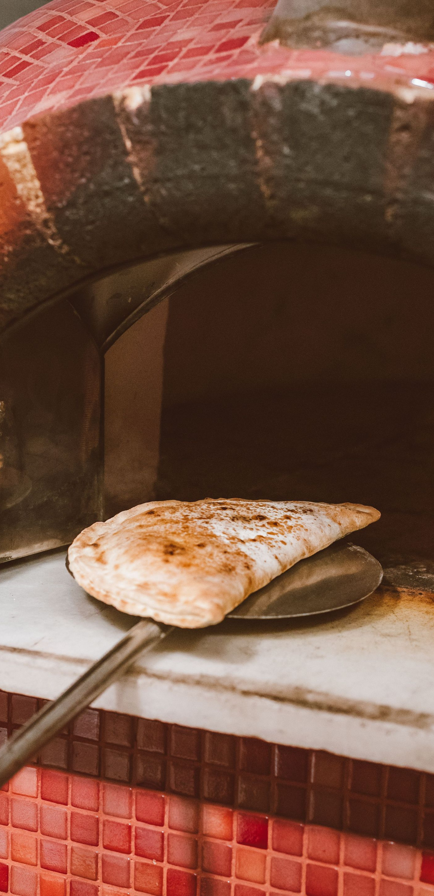 Pizza being removed from a wood-fired oven with a metal peel, red mosaic tiles visible