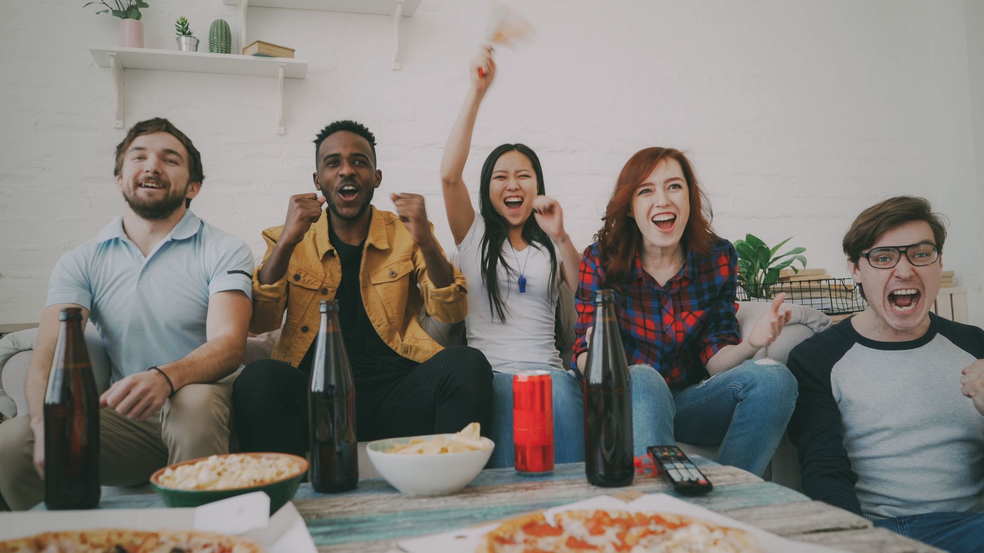 Five friends cheering on a couch with snacks and drinks in a bright living room