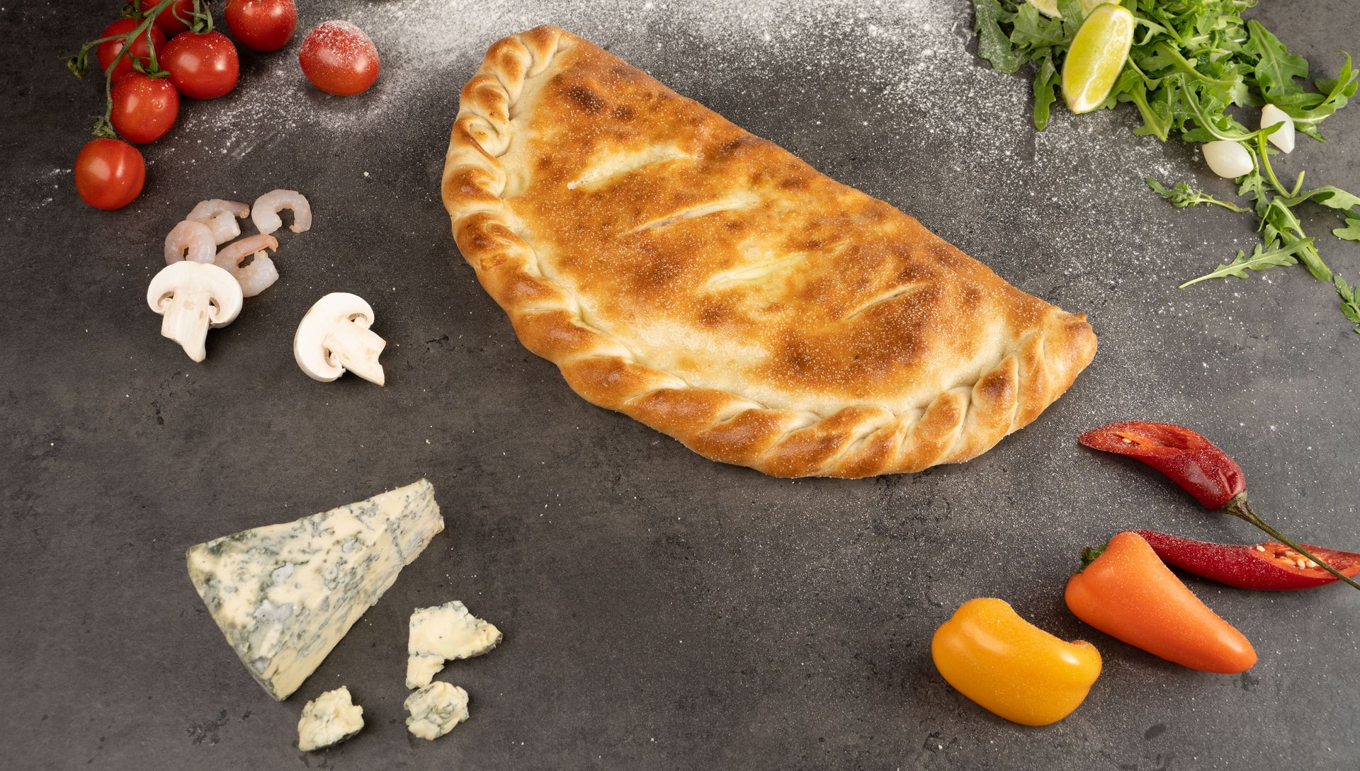 Golden-brown baked calzone on a dark surface, surrounded by mushrooms, tomatoes, peppers, herbs, and cheese