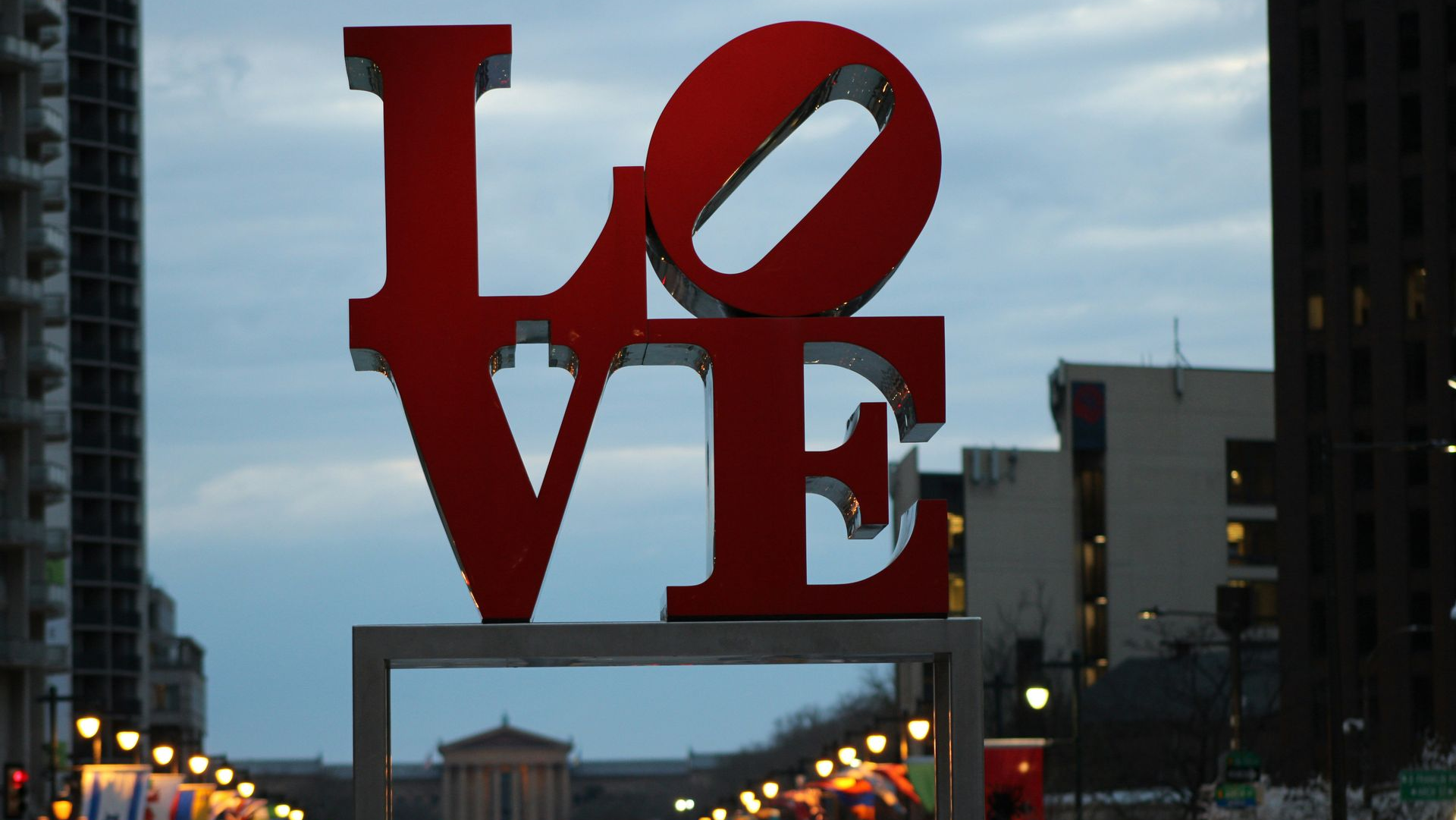The iconic red LOVE sculpture stands in an urban setting, with city buildings and a distant building in the background.