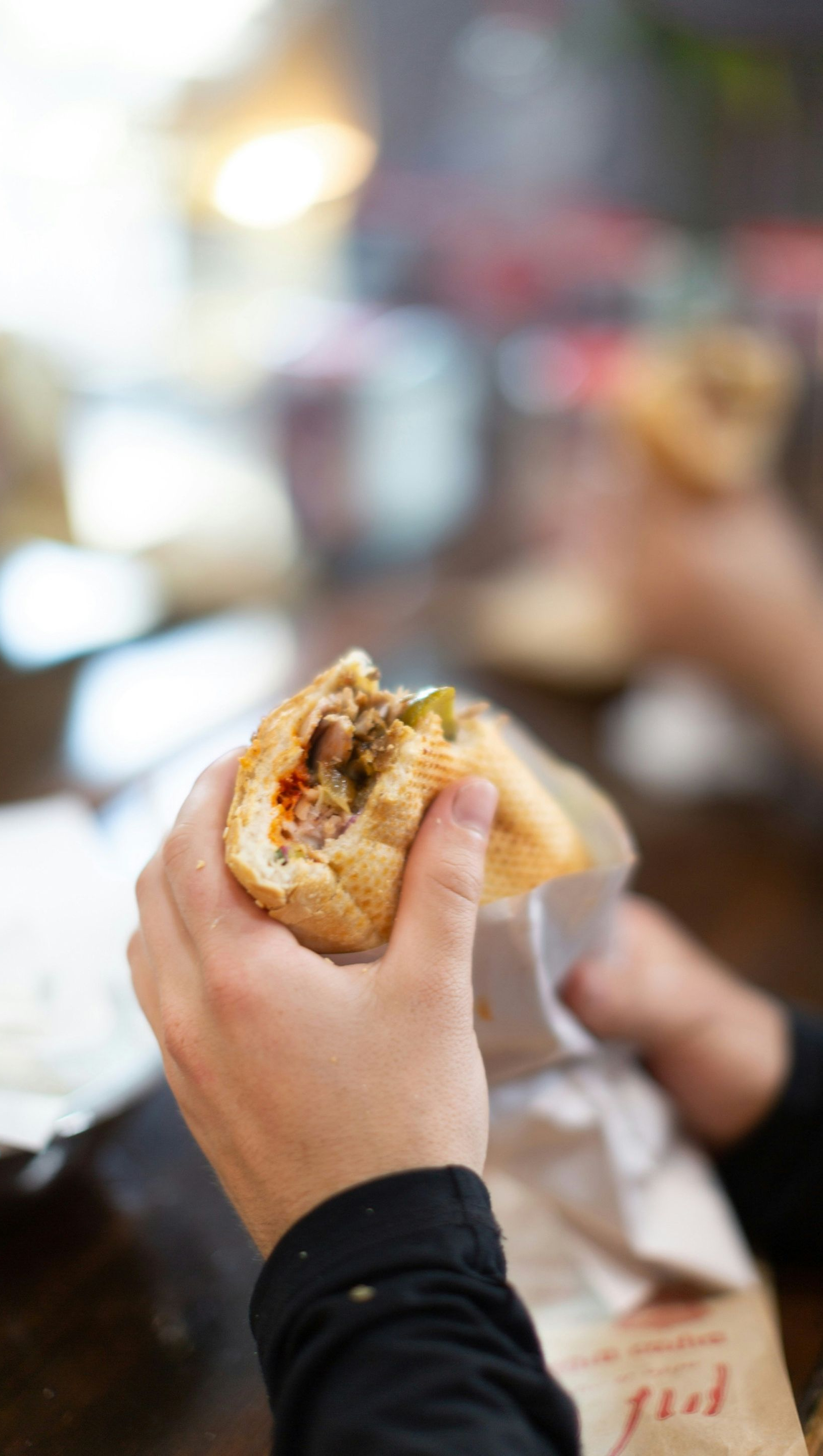 Hands holding a partially eaten sandwich wrapped in paper at a table.