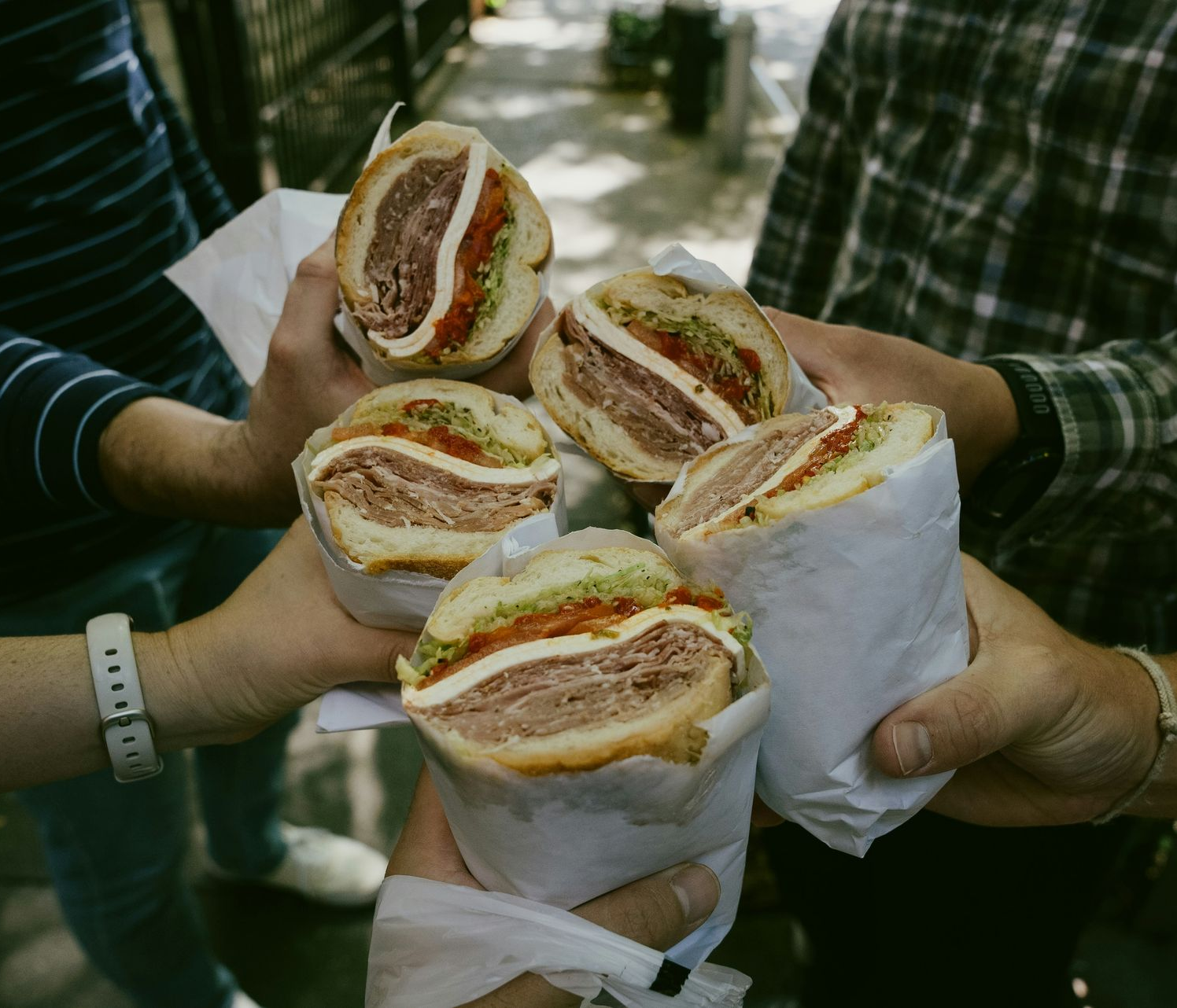 Four people hold up wrapped deli sandwiches together in a toast-like gesture outdoors.
