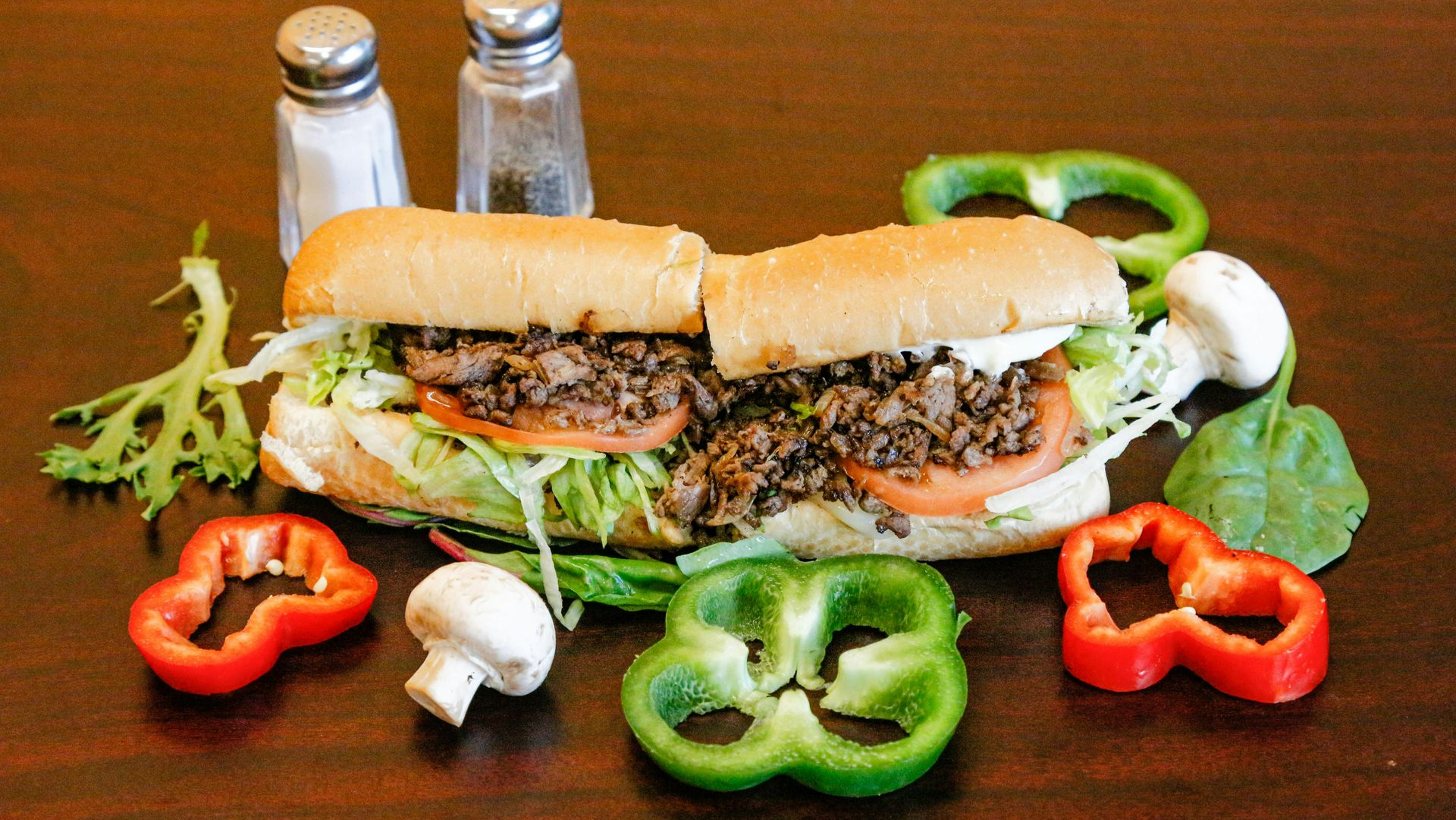 A steak sub sandwich with lettuce and tomato, surrounded by sliced bell peppers, mushrooms, and salt and pepper shakers.
