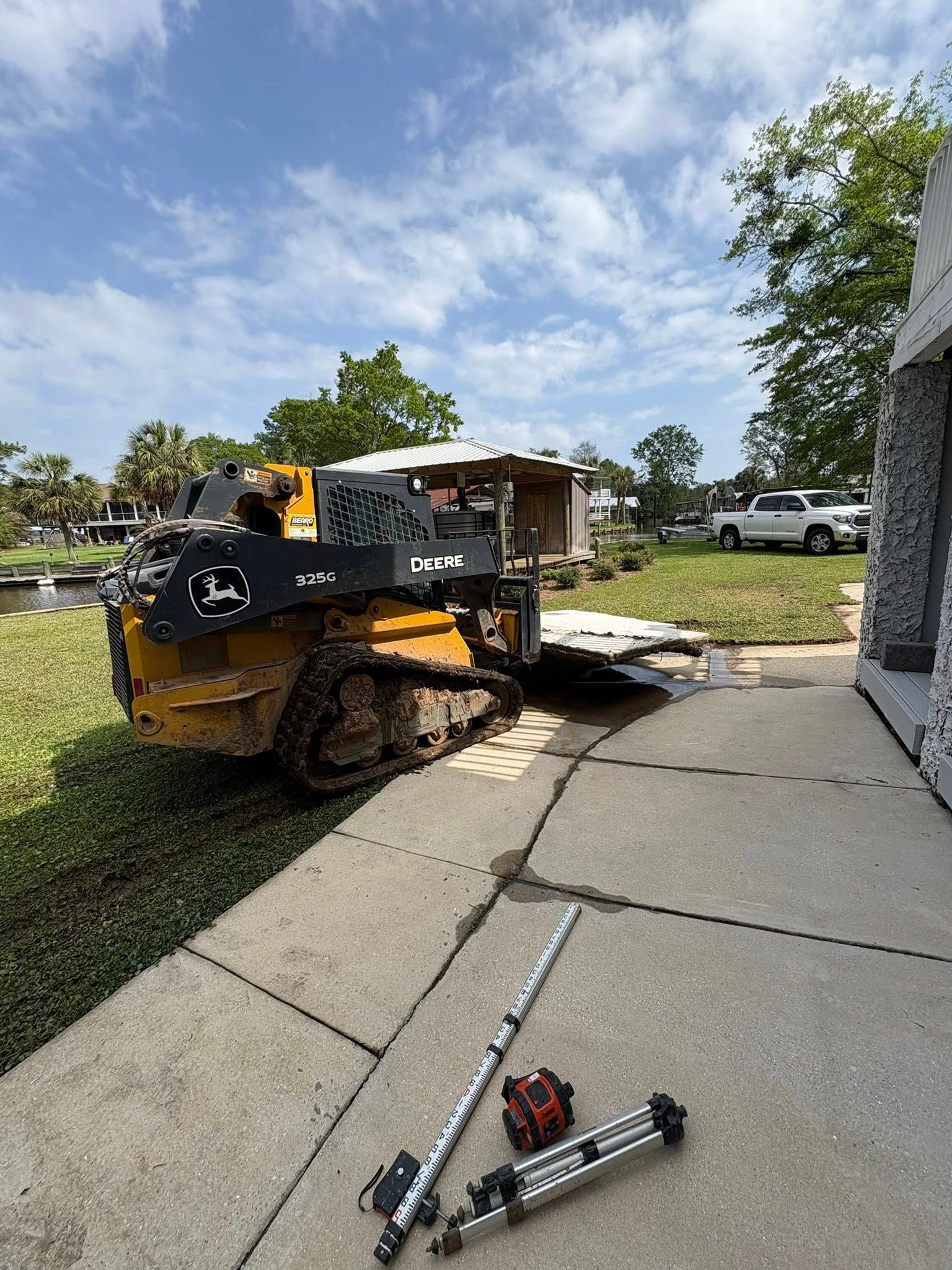 A yellow skid-steer loader sits on grass near a concrete walkway with construction tools lying on the ground.