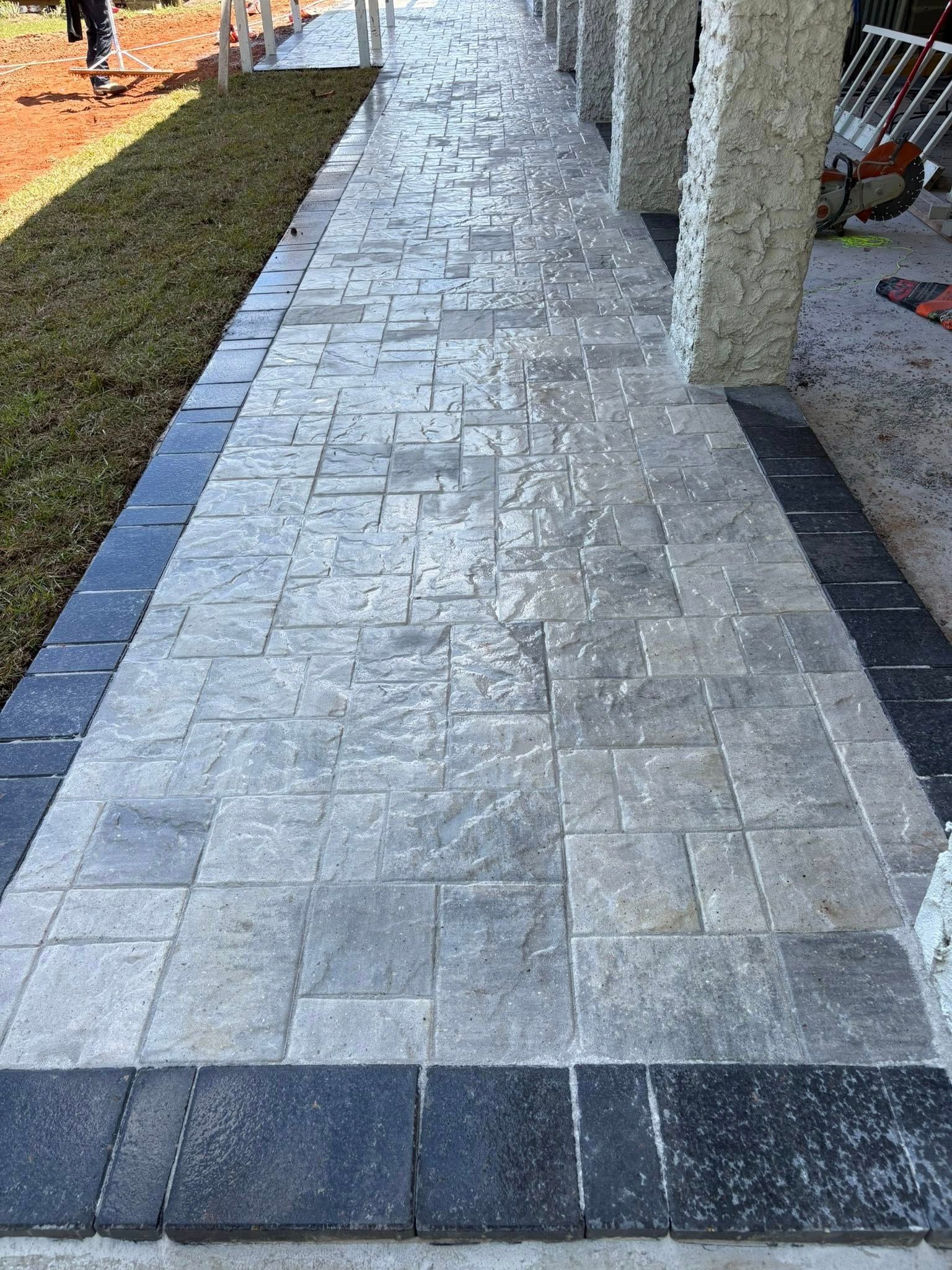 A textured stone walkway featuring a light gray paver pattern bordered by dark, charcoal-colored rectangular tiles.