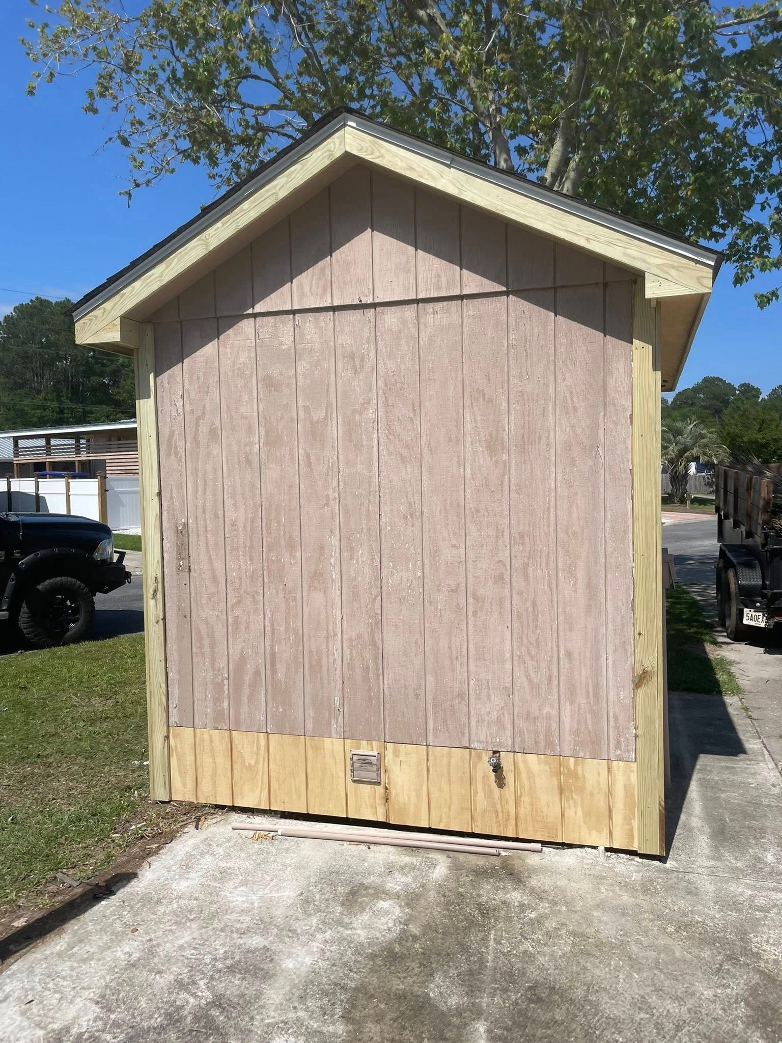 A small shed with weathered brown wood siding and new light-colored wood trim along the base, set on a concrete driveway.