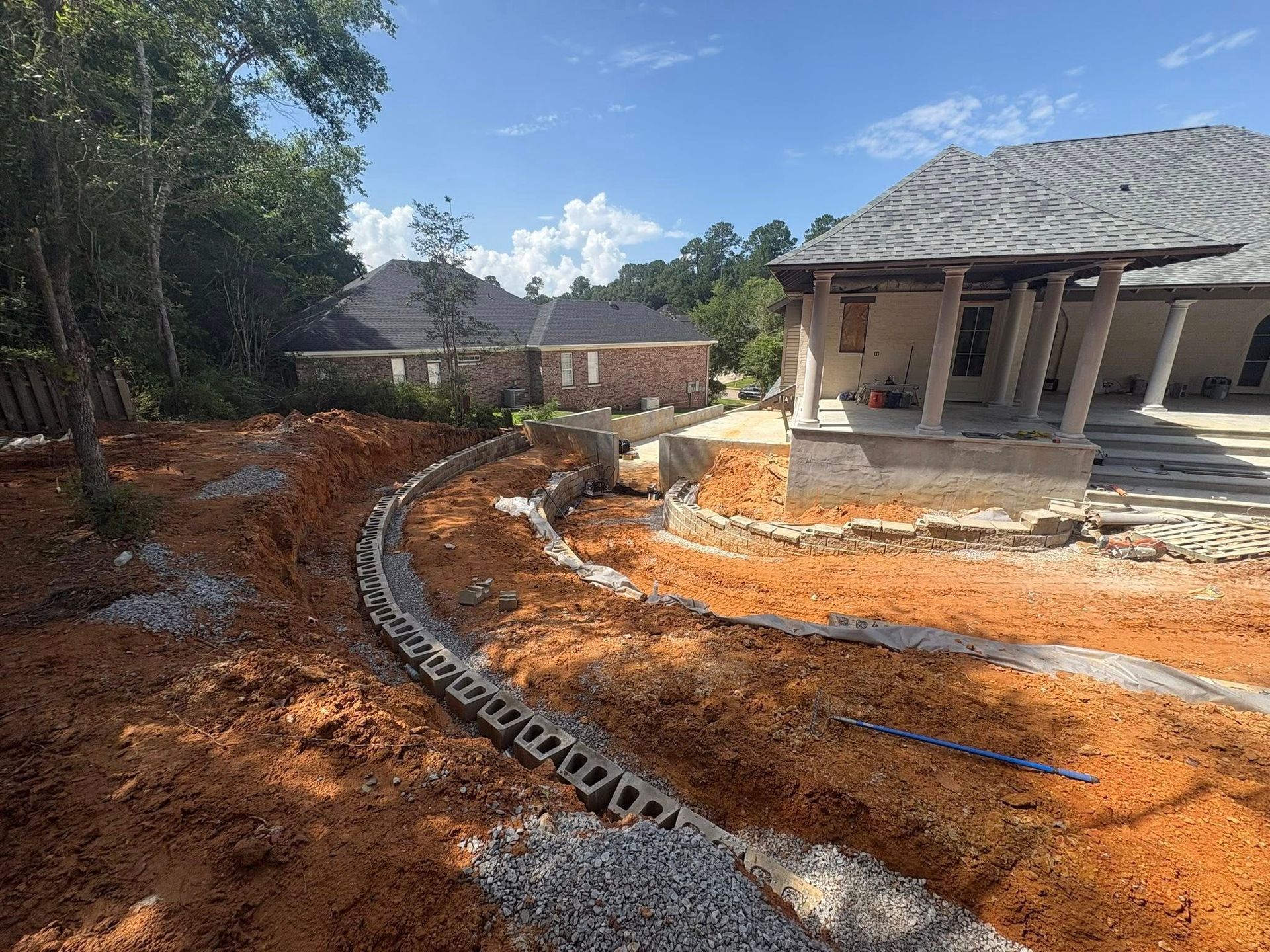 A curved concrete block retaining wall under construction in a reddish-dirt backyard next to a house with a porch.