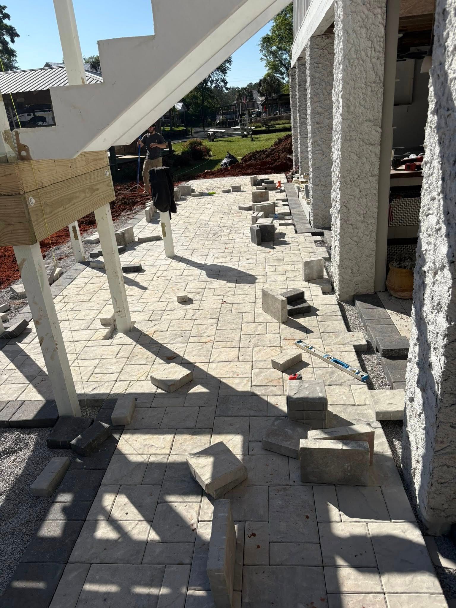 Light-colored stone pavers being laid on a ground-level patio beneath an outdoor staircase and building structure.
