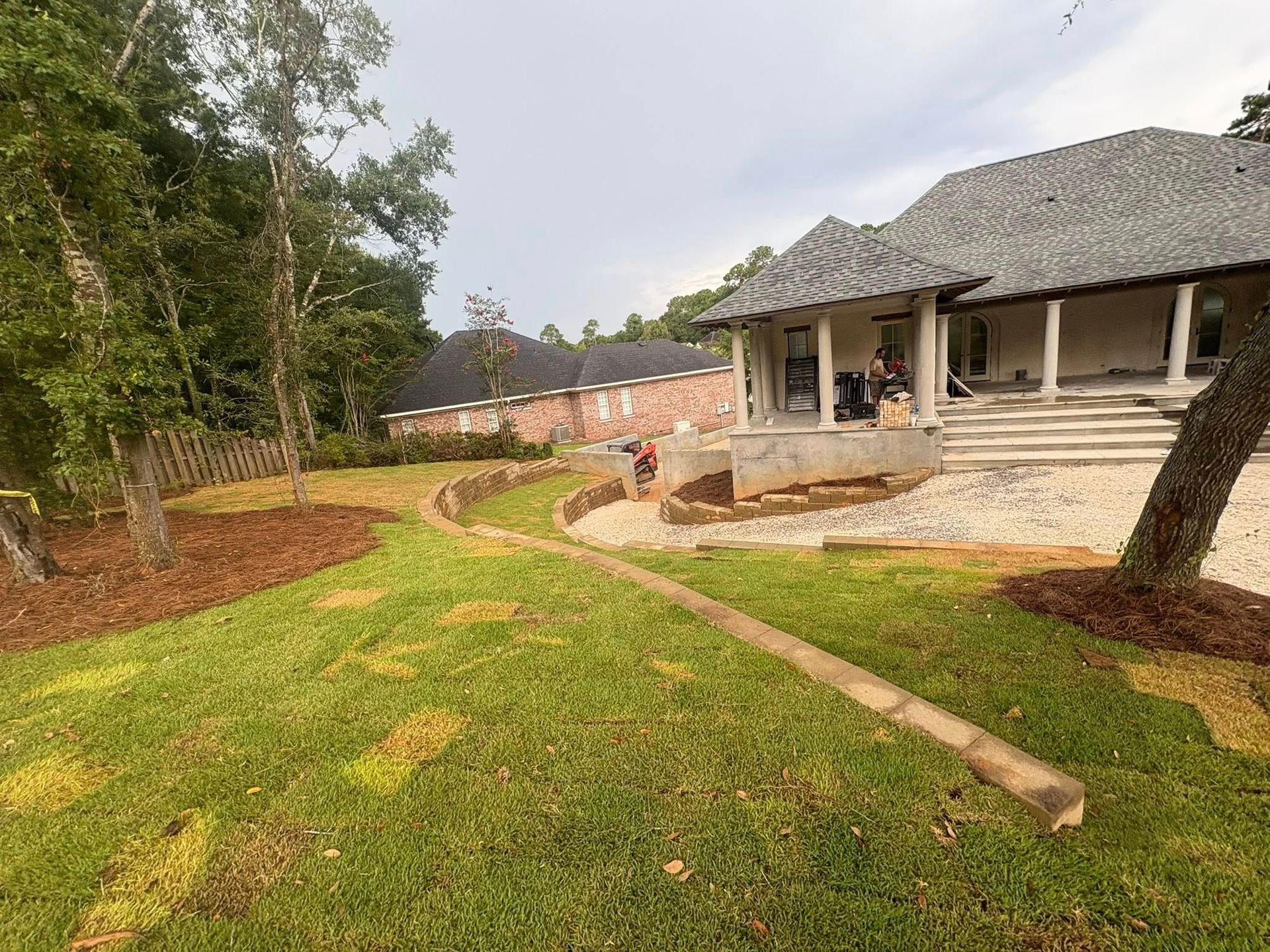 A grassy lawn leads toward a multi-story home under construction with a stone-colored patio and a gravel driveway.