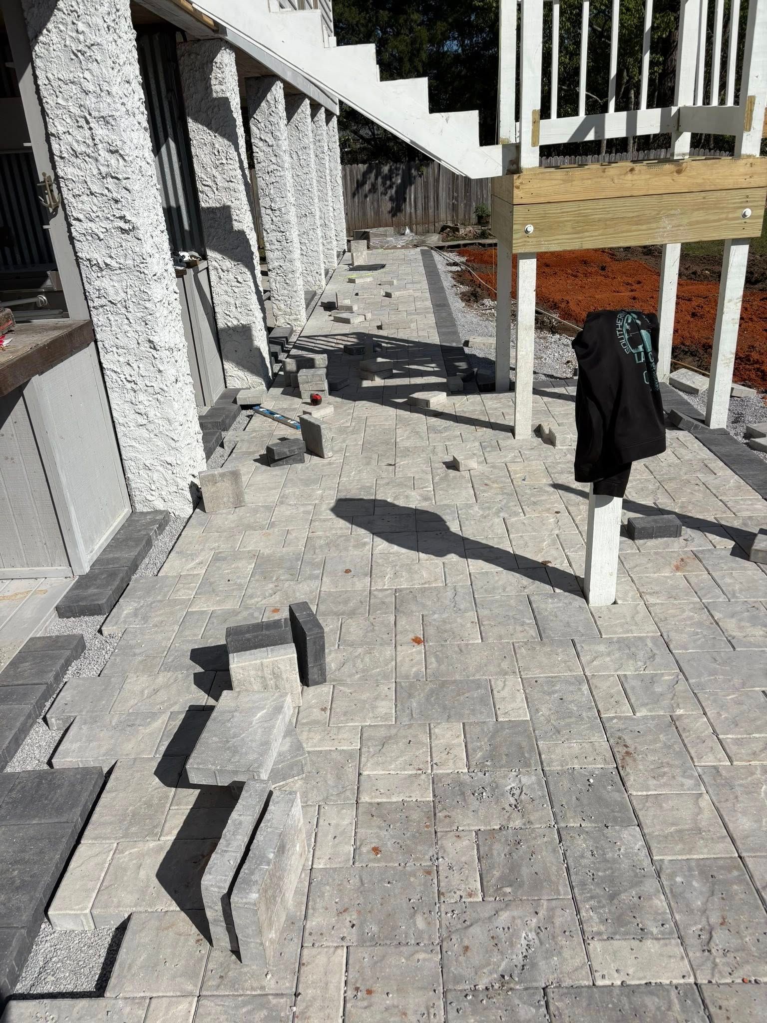 Patio pavers being installed alongside a house foundation under a wooden deck with a jacket hanging on a post.