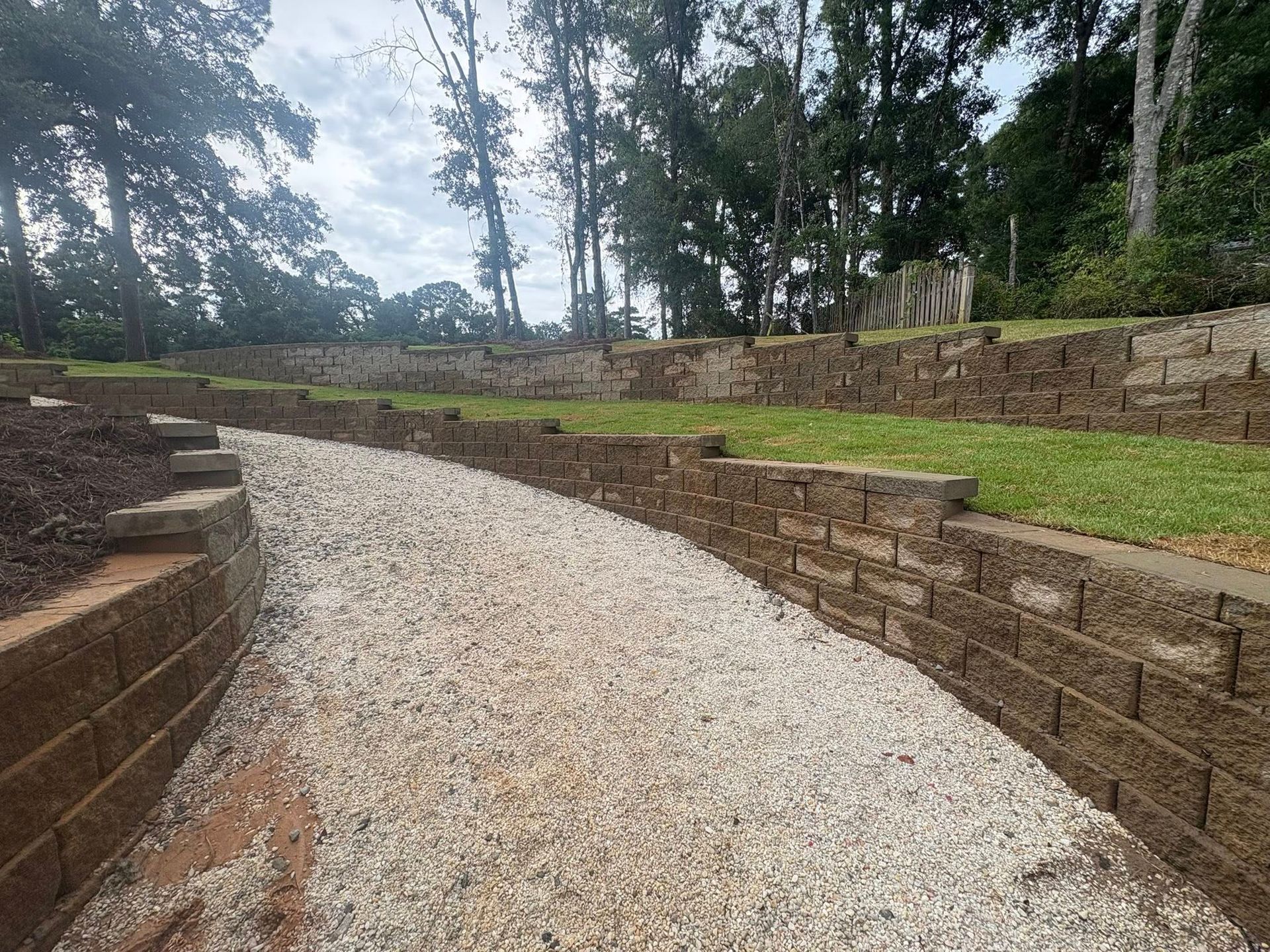 A gravel path curves between tiered, brown stone retaining walls and grassy slopes in a wooded outdoor setting.