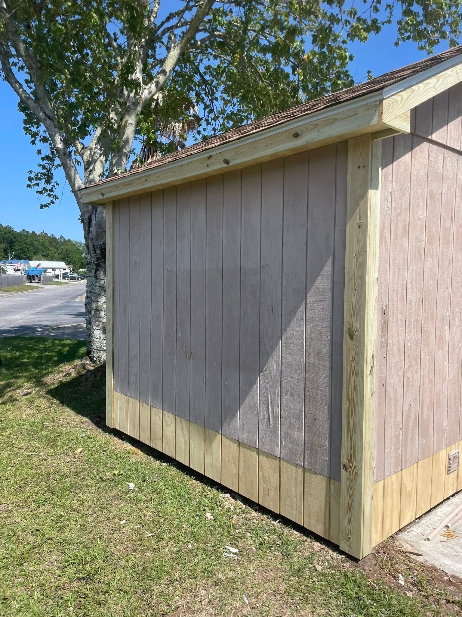 A side view of a small shed with tan vertical siding and new, light-colored wooden trim, set outdoors on a sunny day.