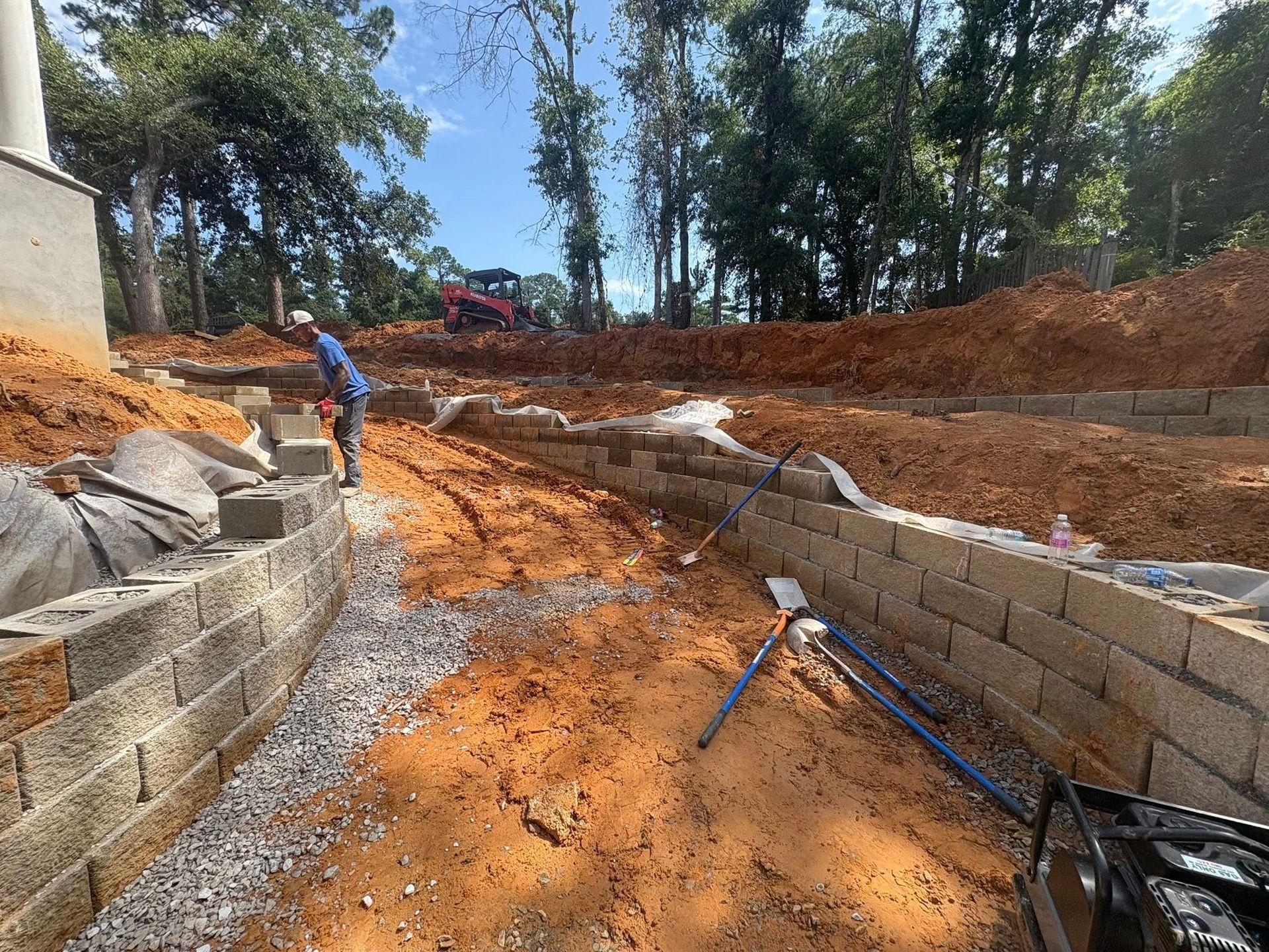A person builds a curved retaining wall with concrete blocks and gravel in an outdoor yard with red soil and trees.