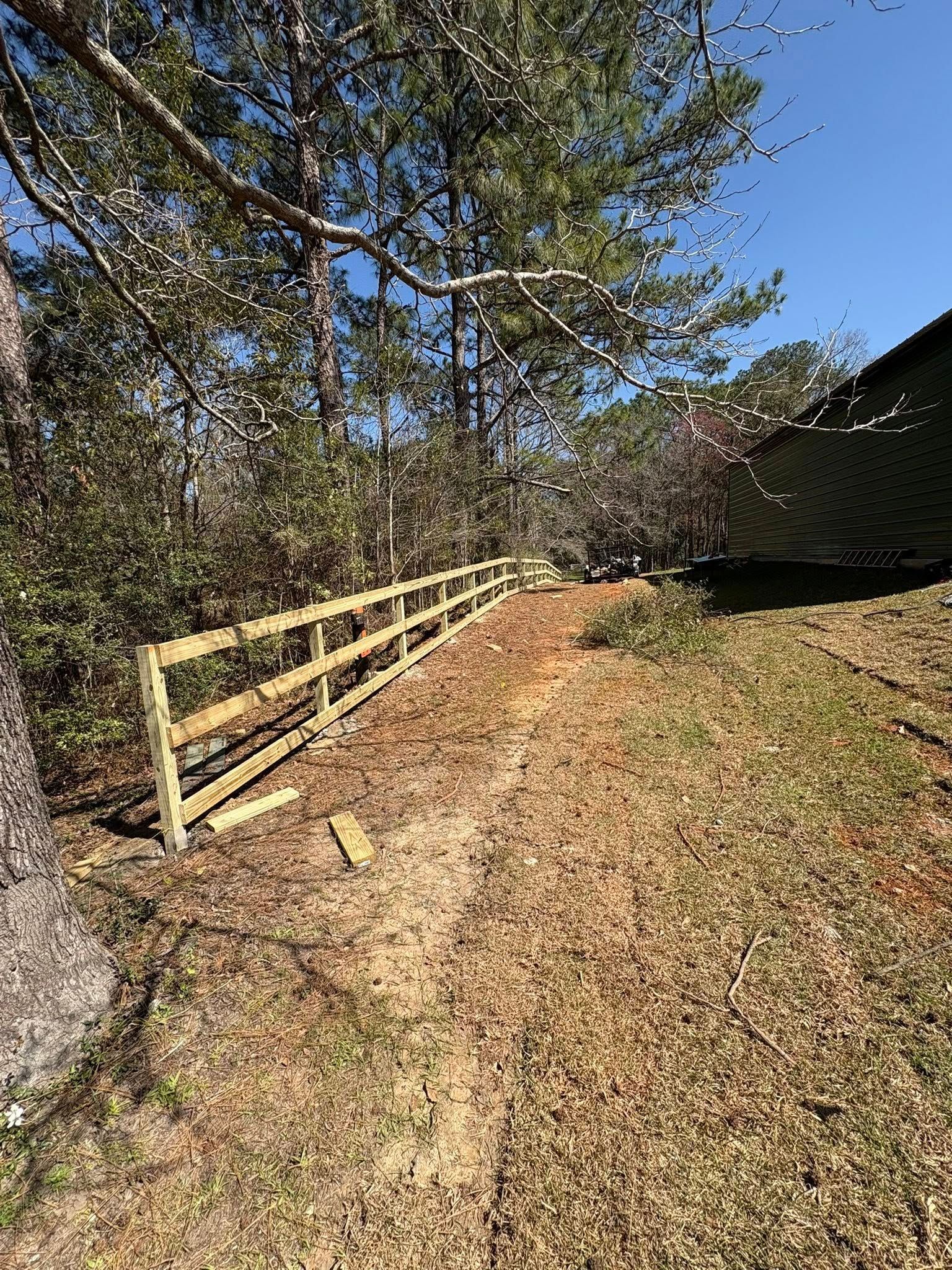 A newly installed wooden three-rail fence runs along a dirt path bordering a wooded area under a clear blue sky.