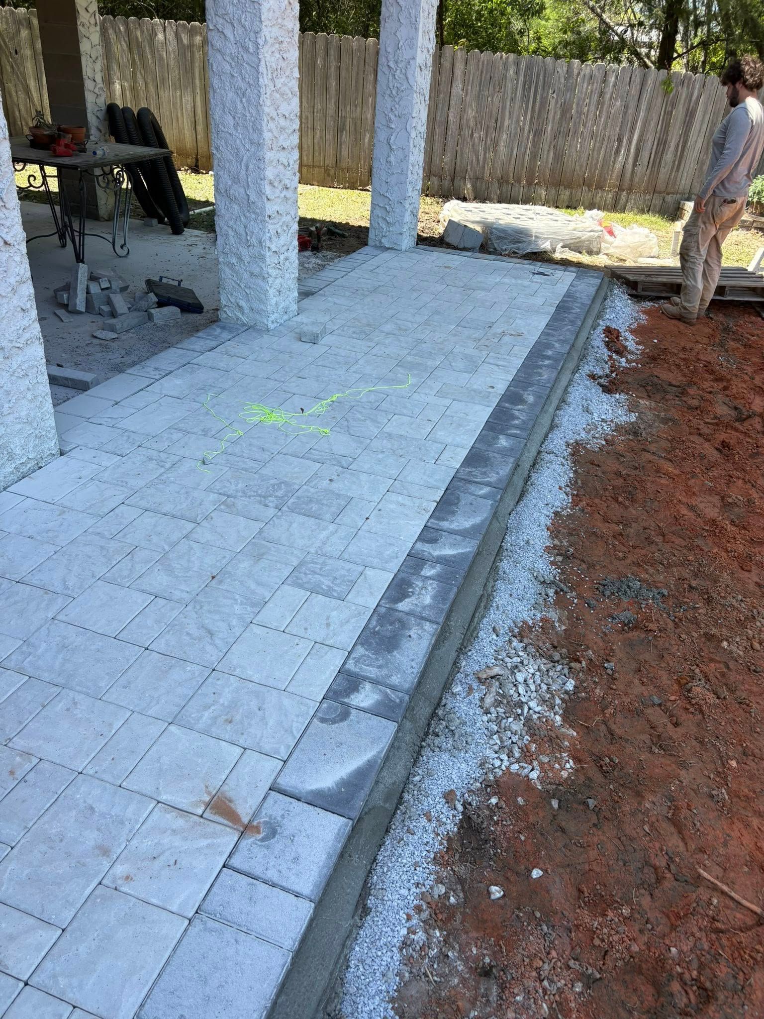 A partially finished patio with light-colored pavers and a dark border, with a person standing on the dirt area nearby.