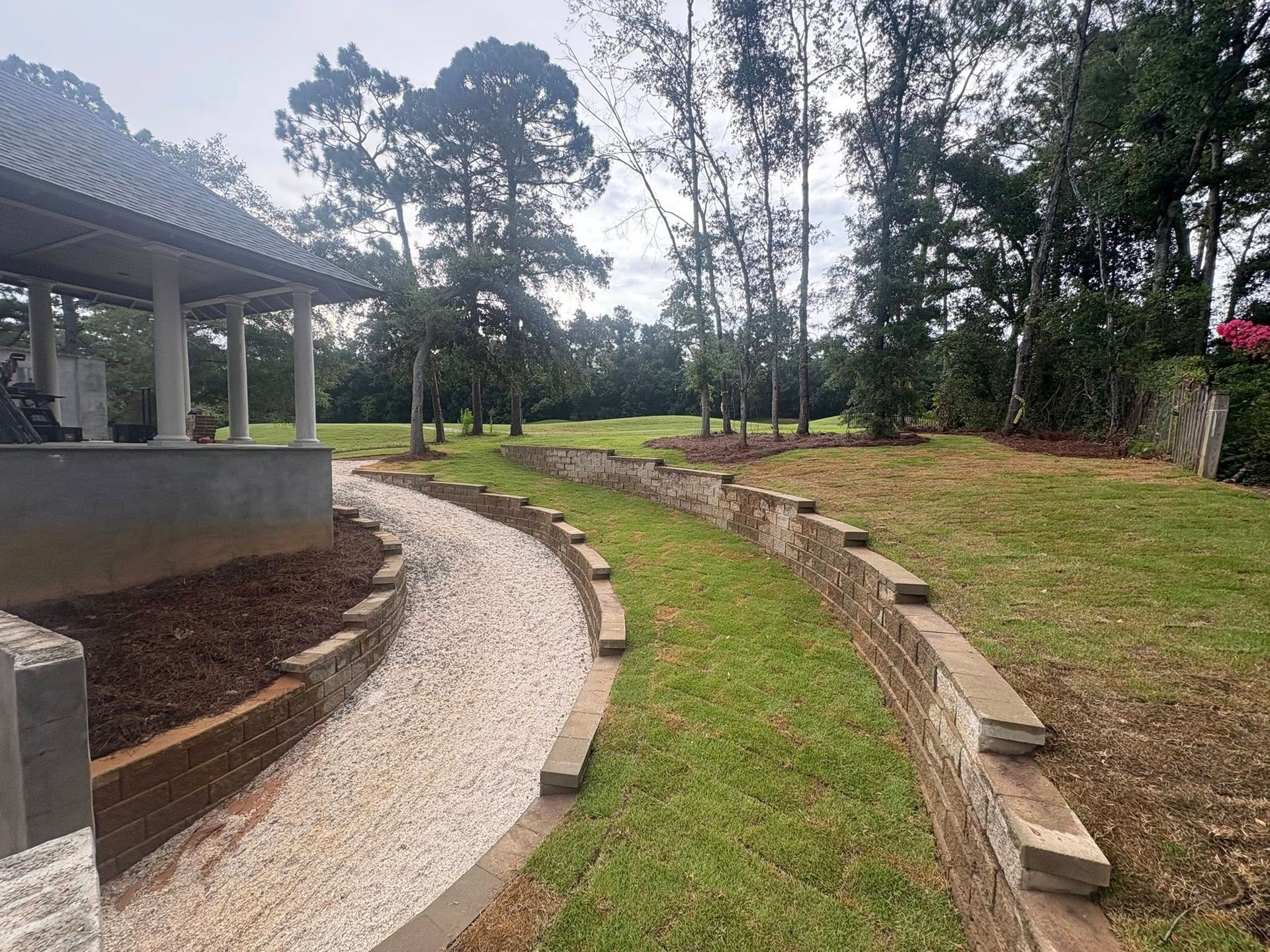 A gravel path winds between a tiered stone retaining wall and a covered patio area in a landscaped yard.