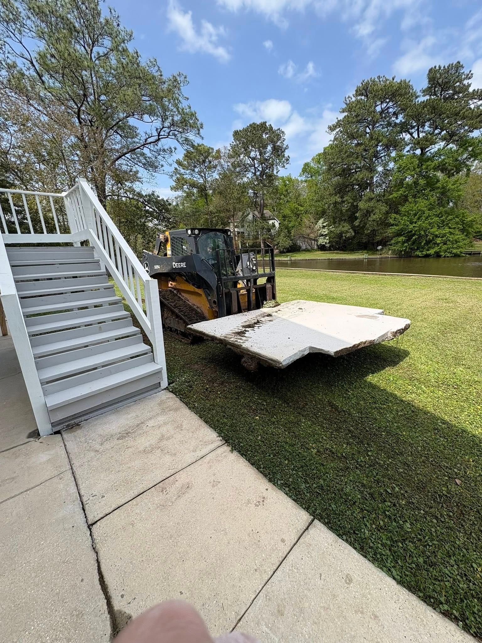 A yellow skid steer loader carries a large concrete slab across a grassy yard near a wooden staircase and a body of water.