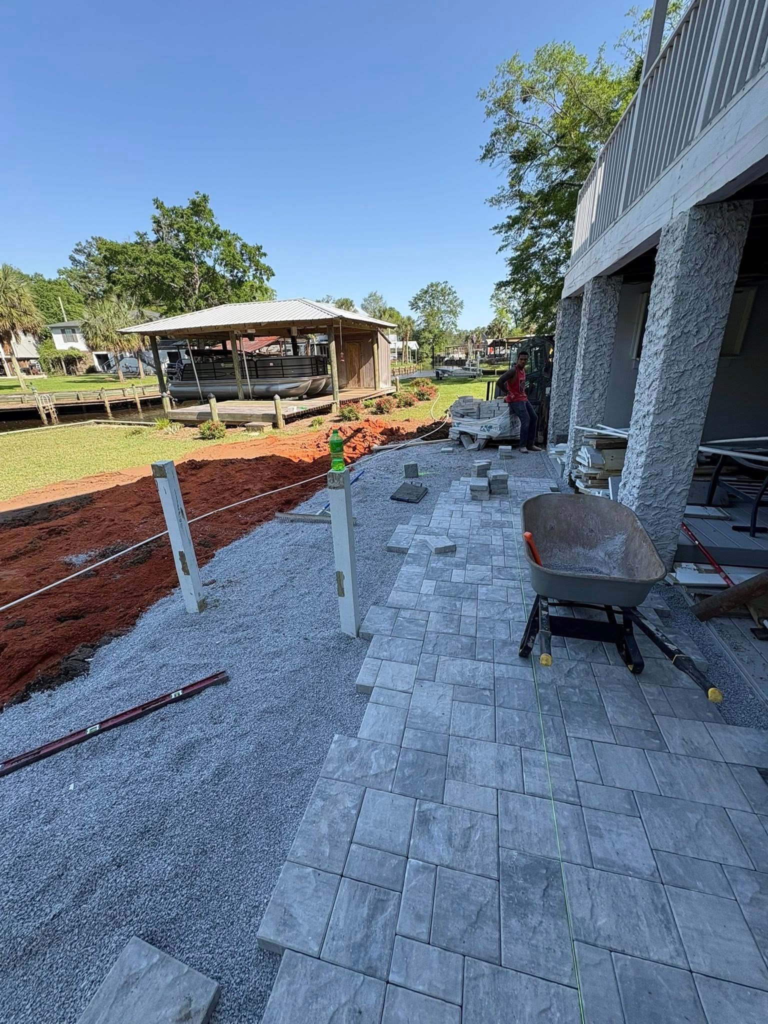 A stone-paved patio under construction next to a house with white textured columns, a gravel border, and a wheelbarrow.