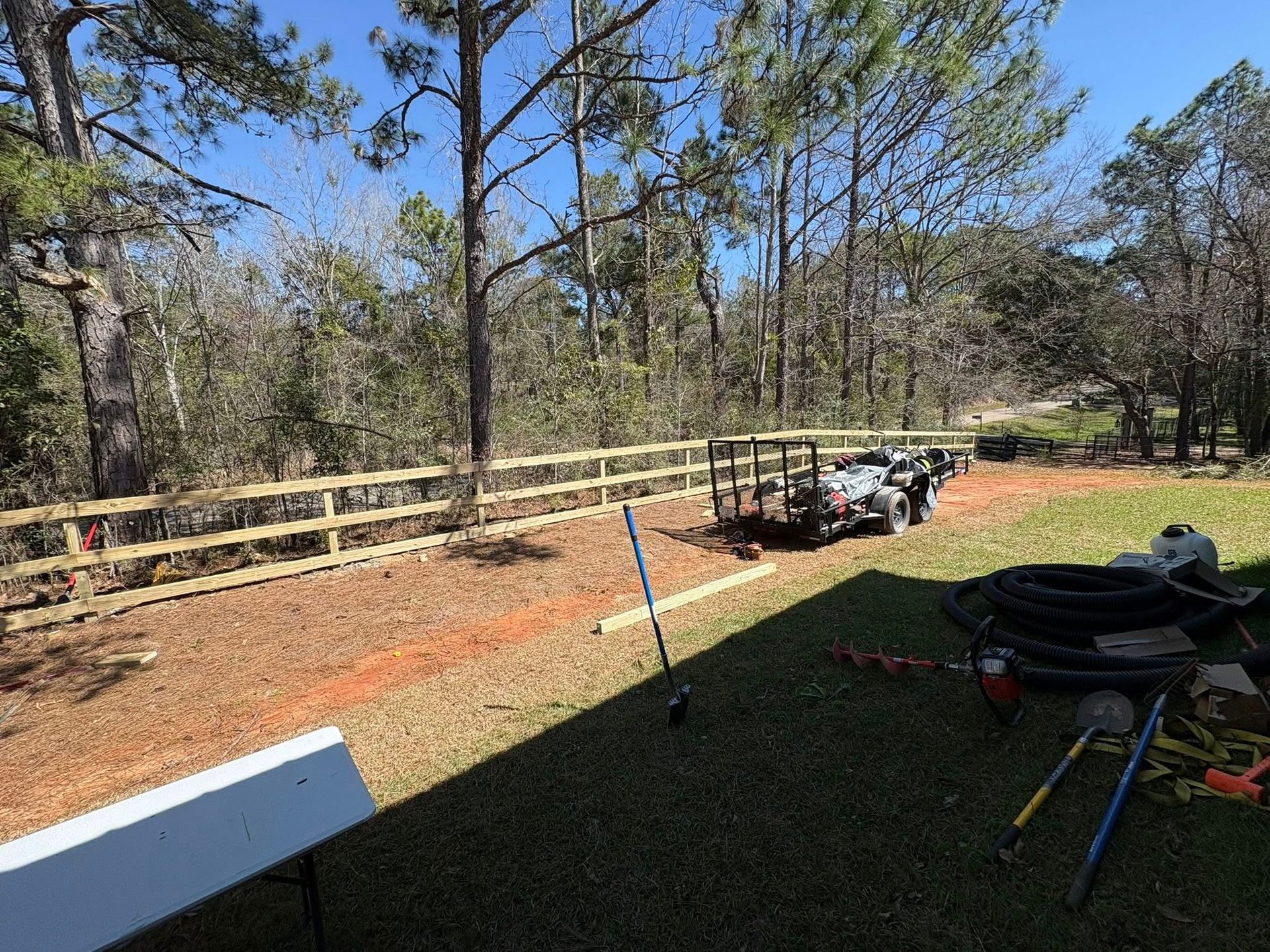 A wooden fence borders a backyard with a clearing, a trailer, tools, and trees under a clear blue sky.