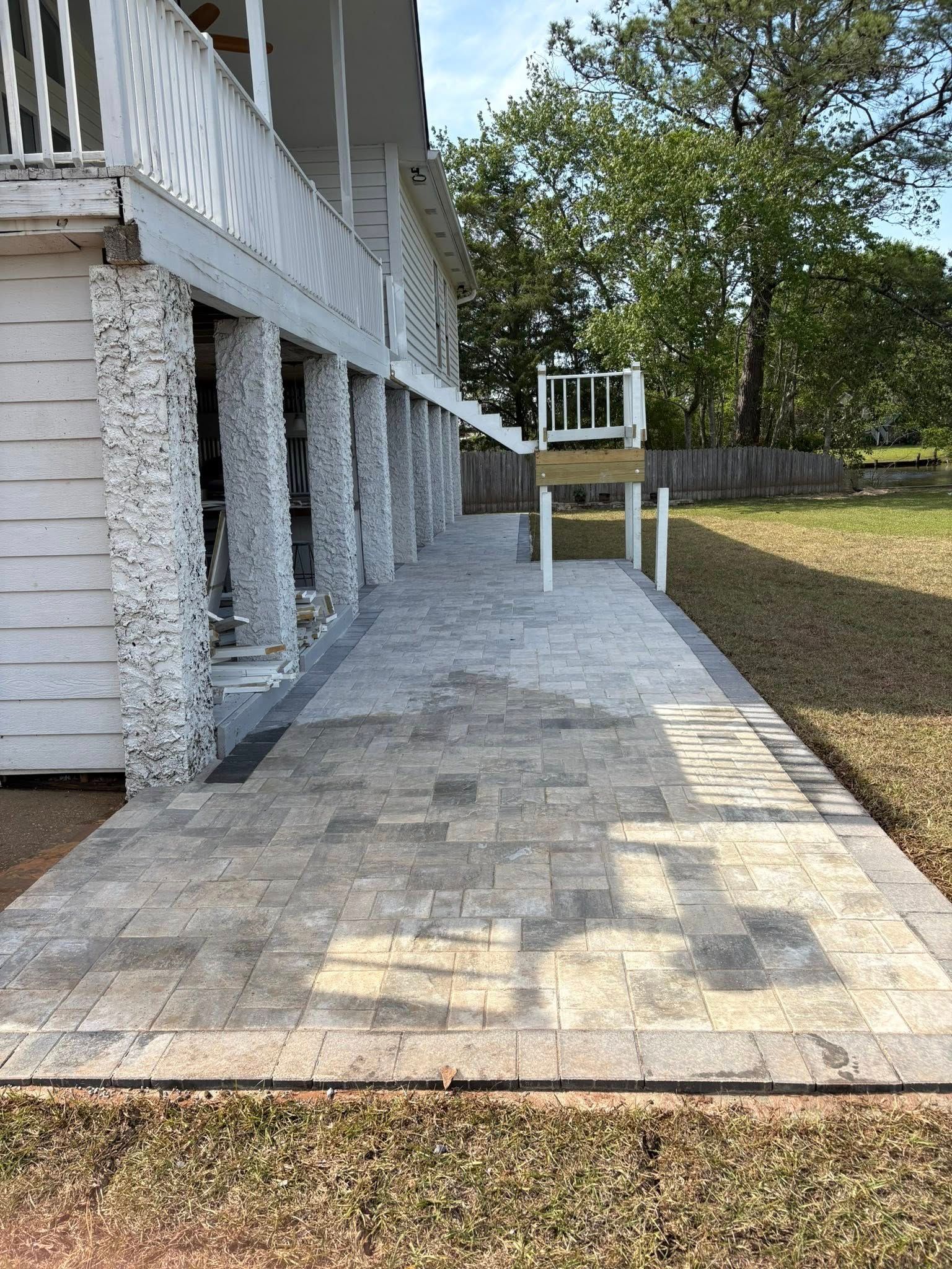 A newly installed stone paver patio walkway alongside a house with white porch pillars and a small wooden play structure.