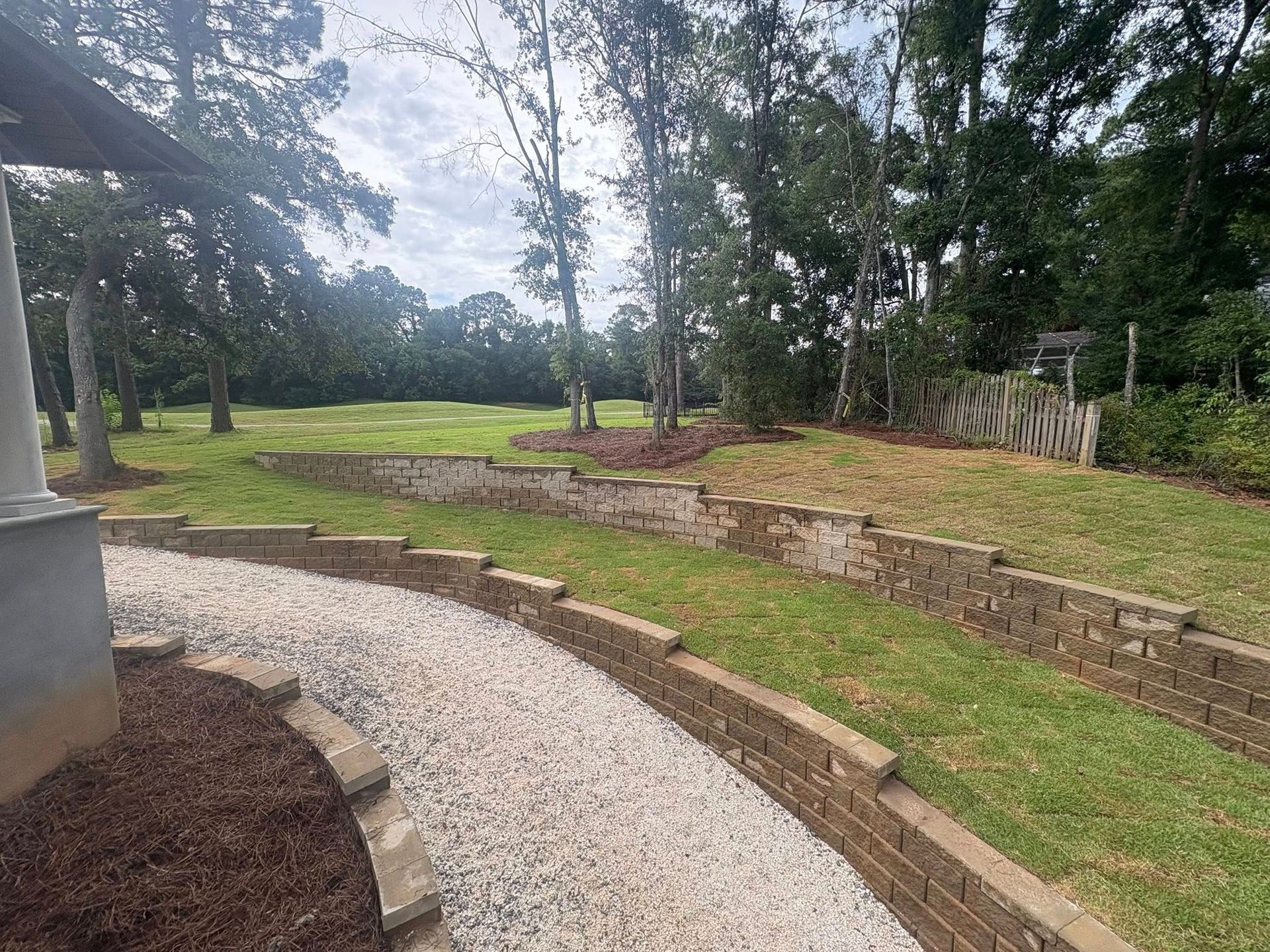 A curved gravel path leads past a tiered stone retaining wall through a grassy lawn toward a line of trees.