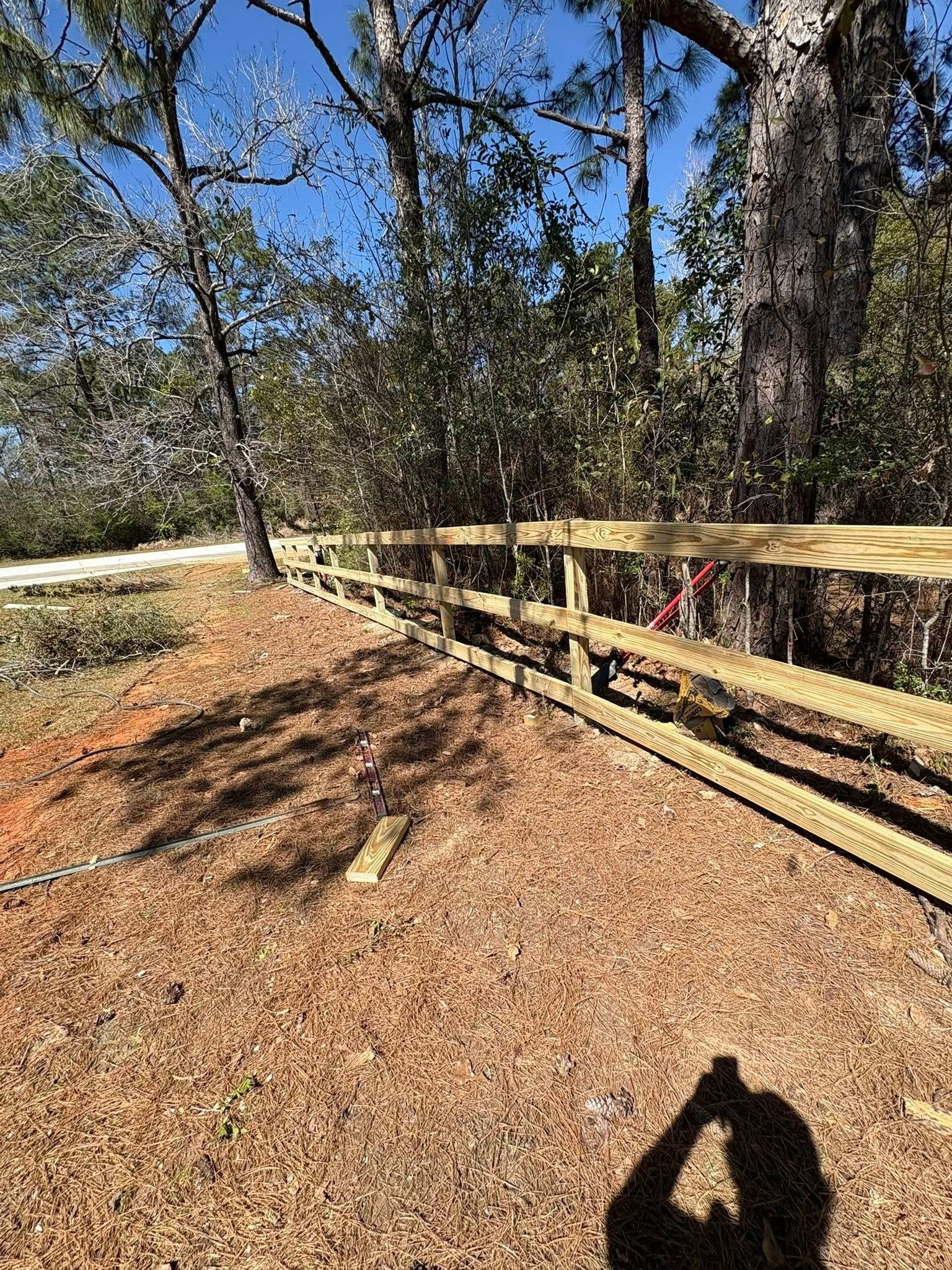 A newly installed wooden split-rail fence runs through a wooded area covered in brown pine needles under a blue sky.
