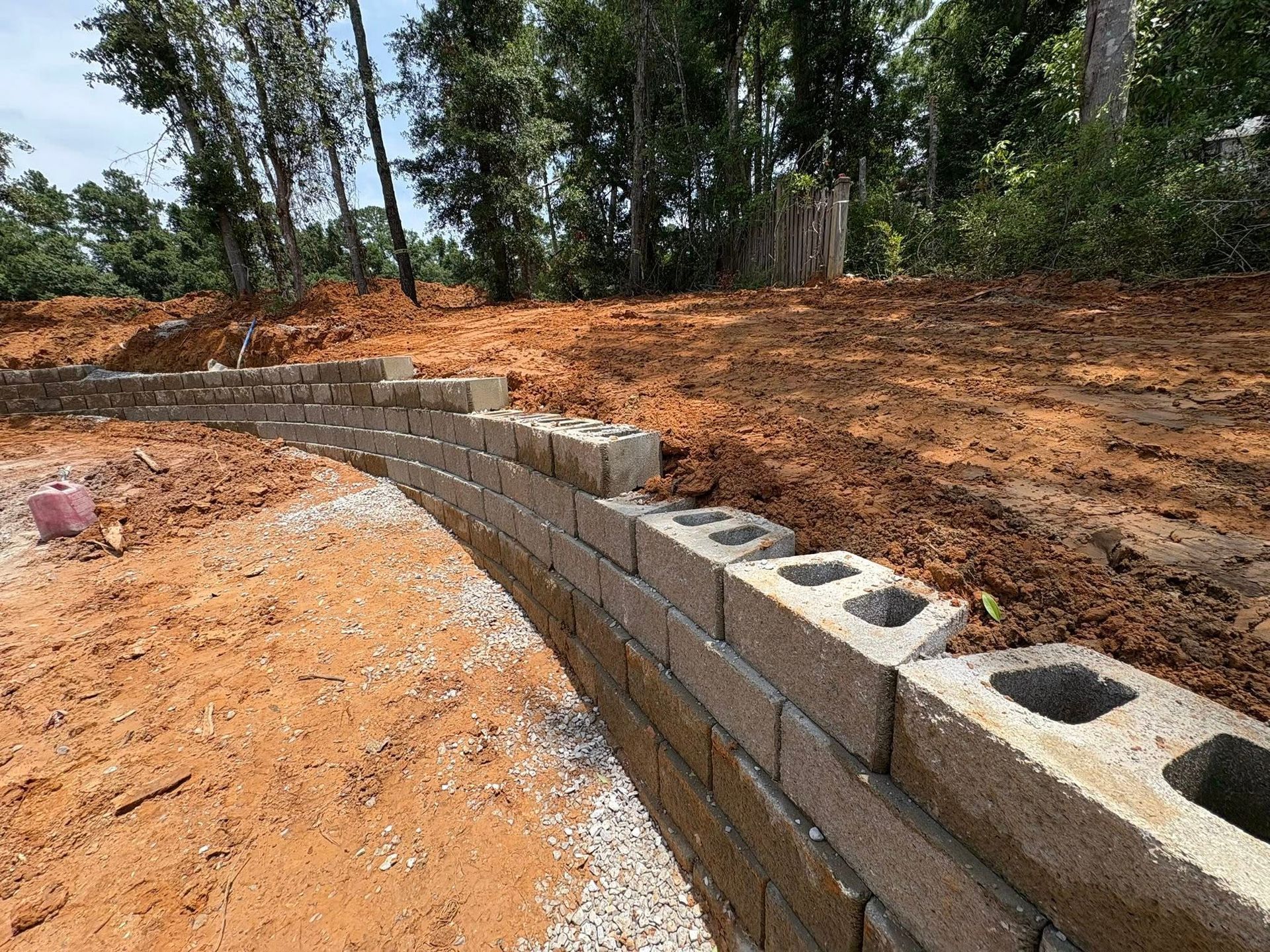 A curved retaining wall made of concrete blocks, partially built on a red-dirt hillside with trees in the background.