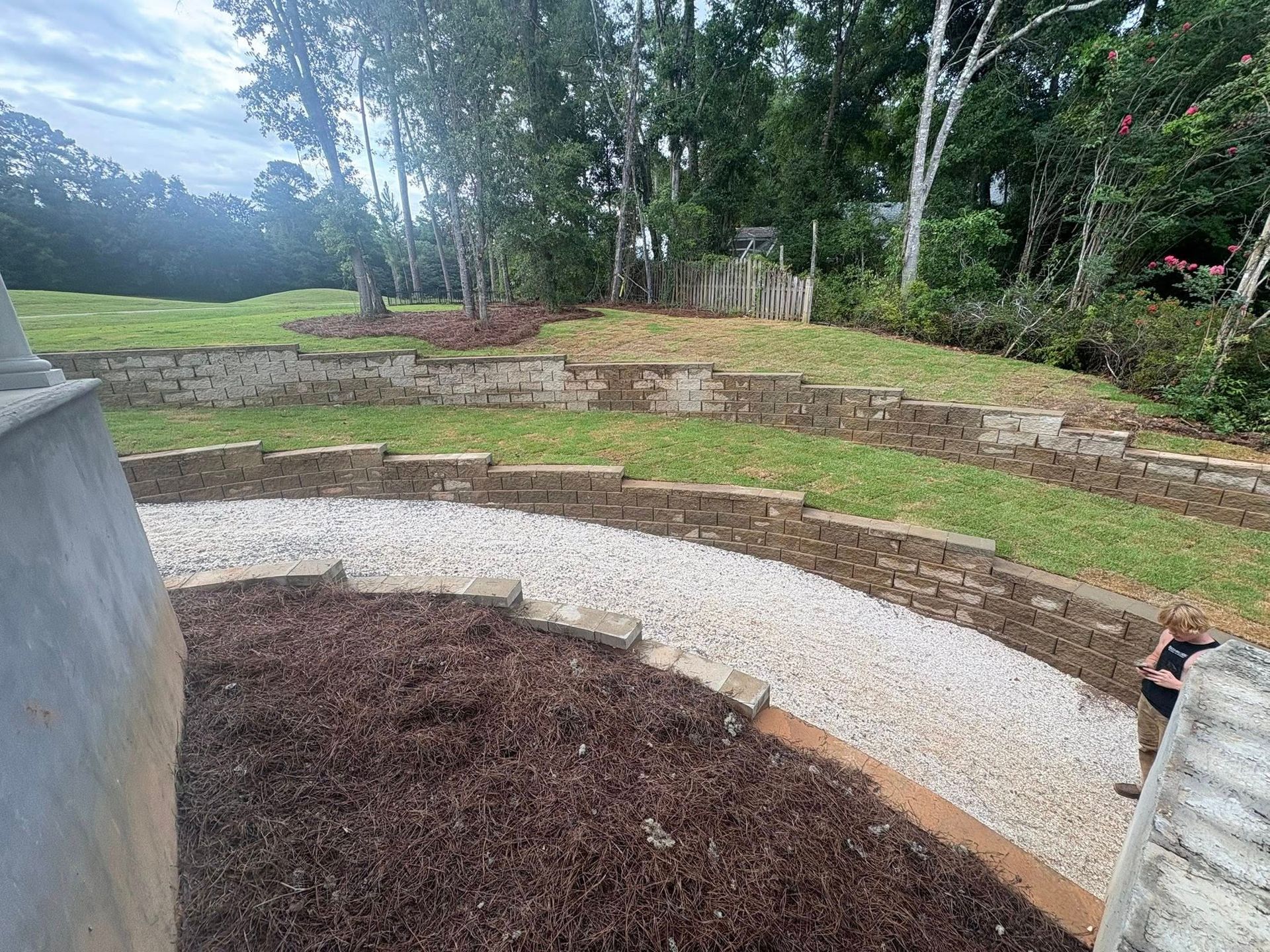 A curved, gravel garden path winds through a landscaped yard with tiered stone retaining walls and mulch beds.
