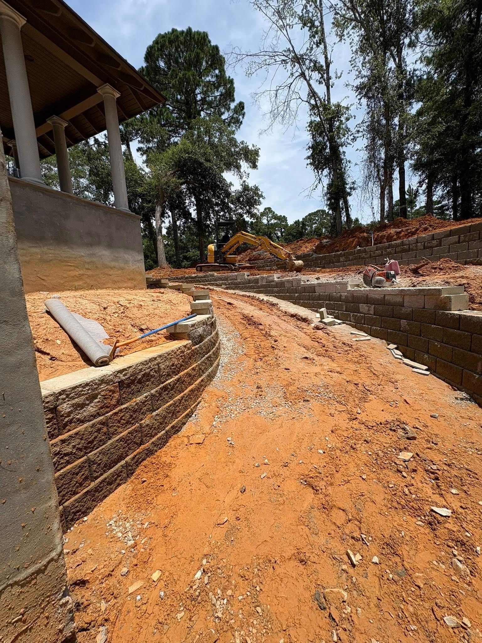 A construction site with a new concrete block retaining wall along a dirt path leading to a house porch under trees.