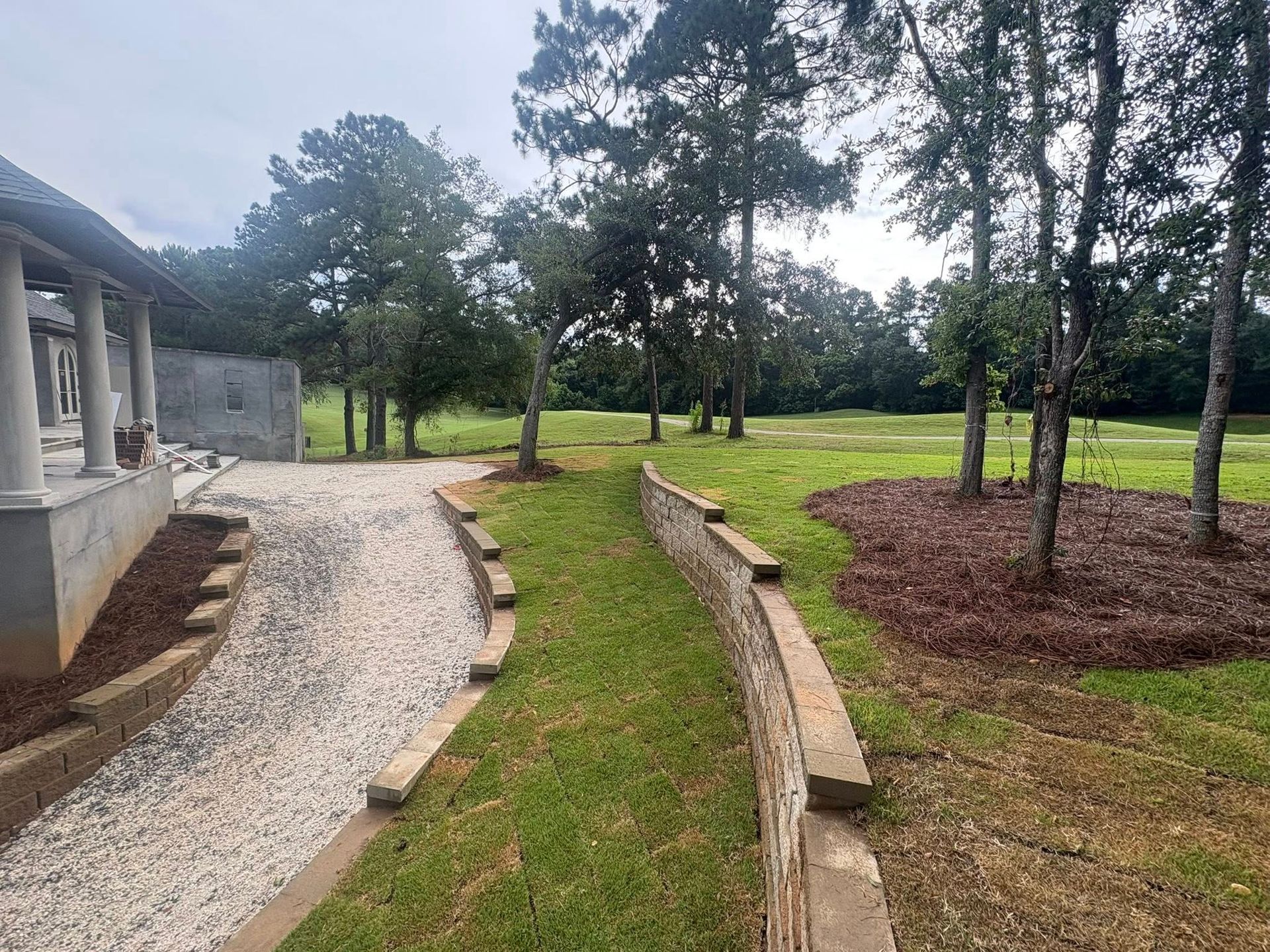 A gravel path winds past a curved stone retaining wall through a grassy yard toward trees and an open field.