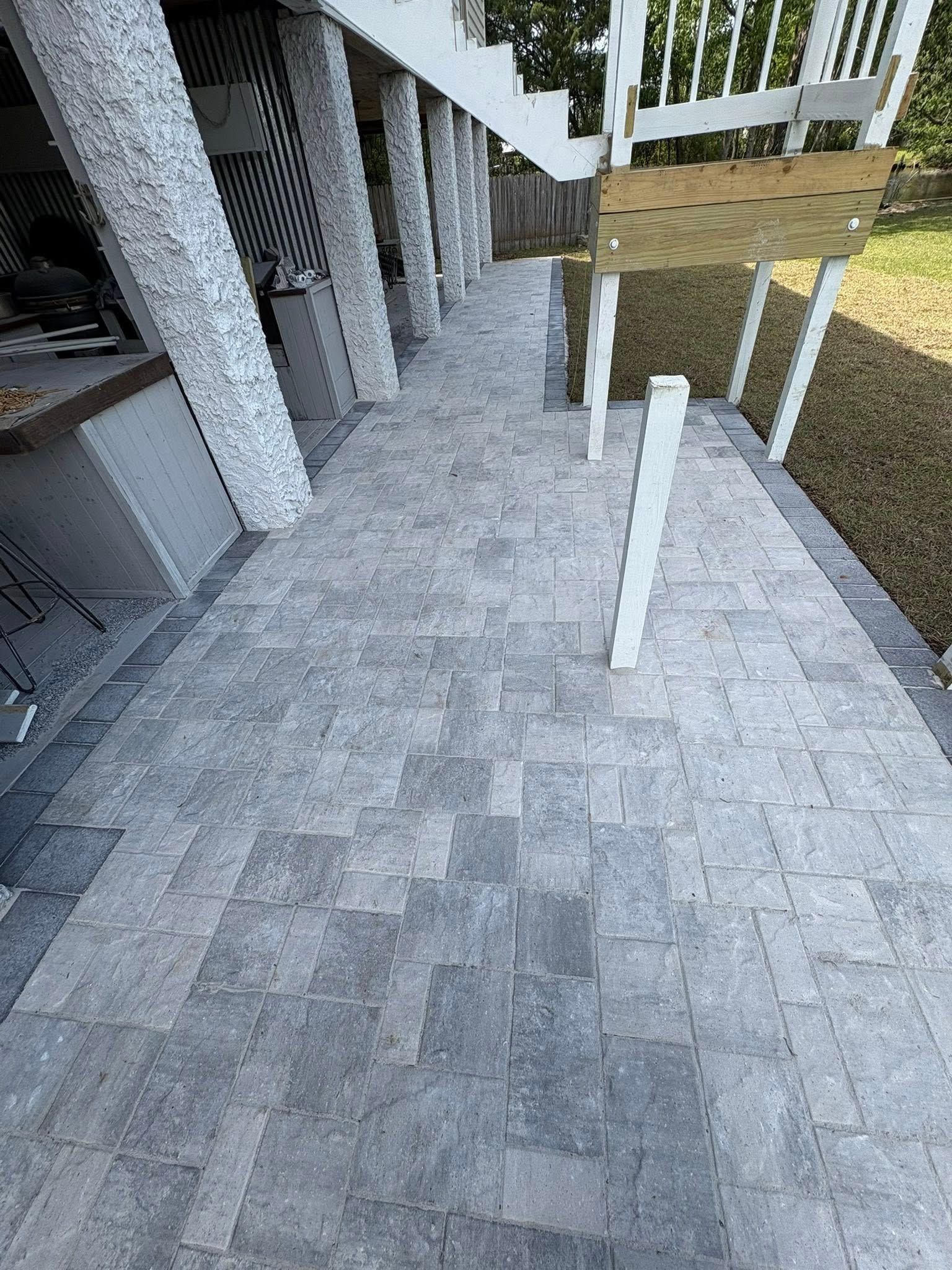 A patio paved with gray rectangular stones, stretching along a house with white support columns and a wooden staircase.