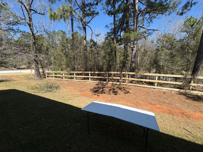 A white folding table sits on a grassy lawn in front of a wooden fence and a forest under a bright blue sky.
