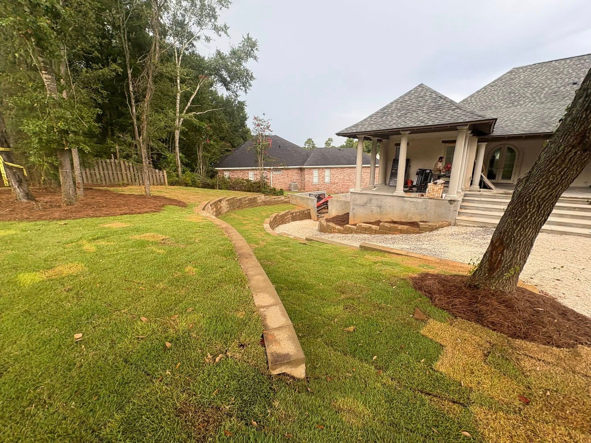 A backyard landscape with a stone retaining wall curving through a lawn, near a large house with a covered patio.