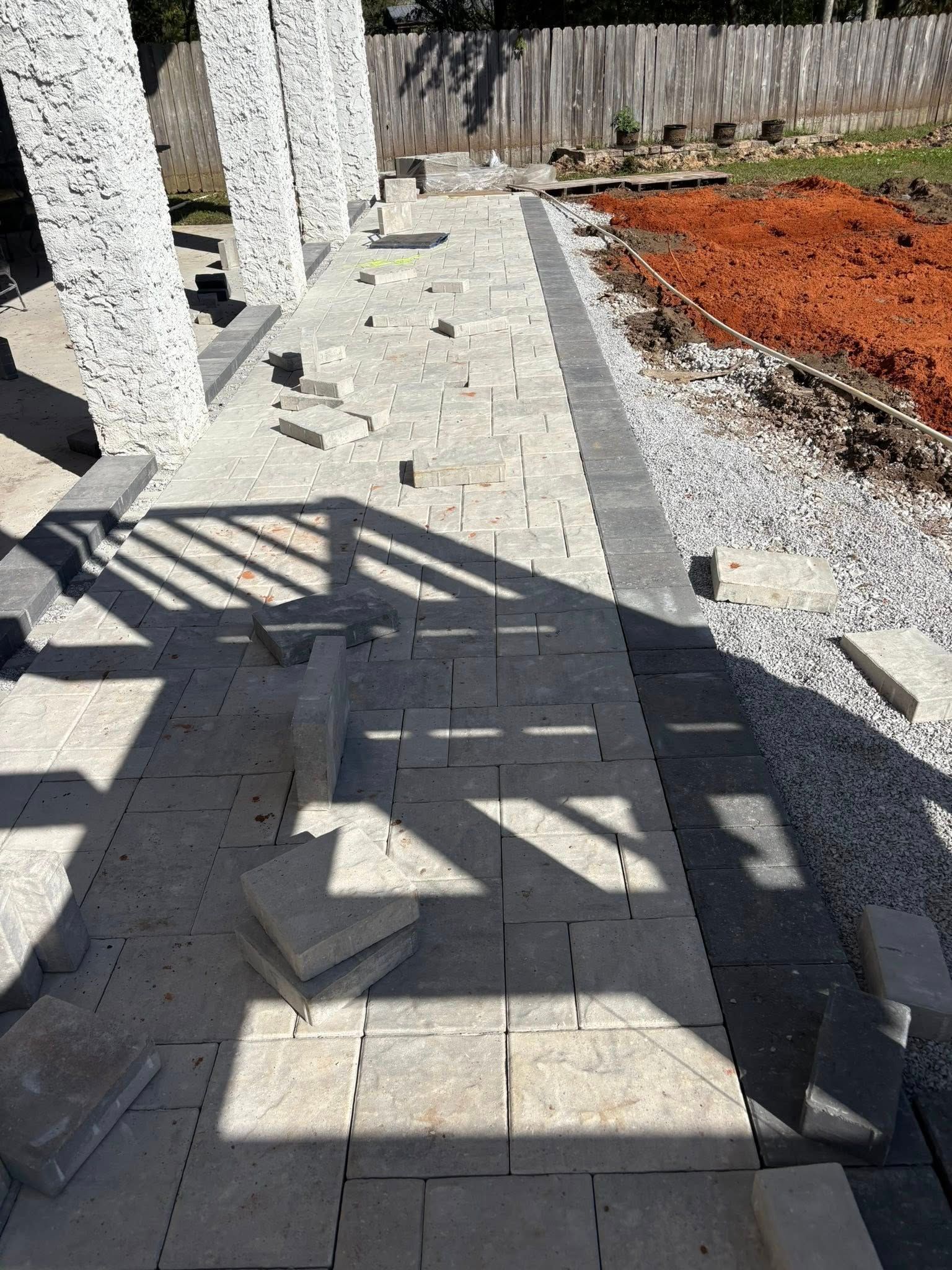 A partially laid stone paver walkway next to white textured pillars and a patch of red soil.